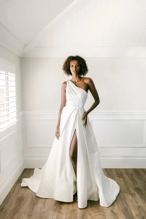 A woman stands indoors wearing a white EB22531-1Z one-shoulder gown with a high slit. She has short curly hair and is posing in a bright room with wood floors and white walls.