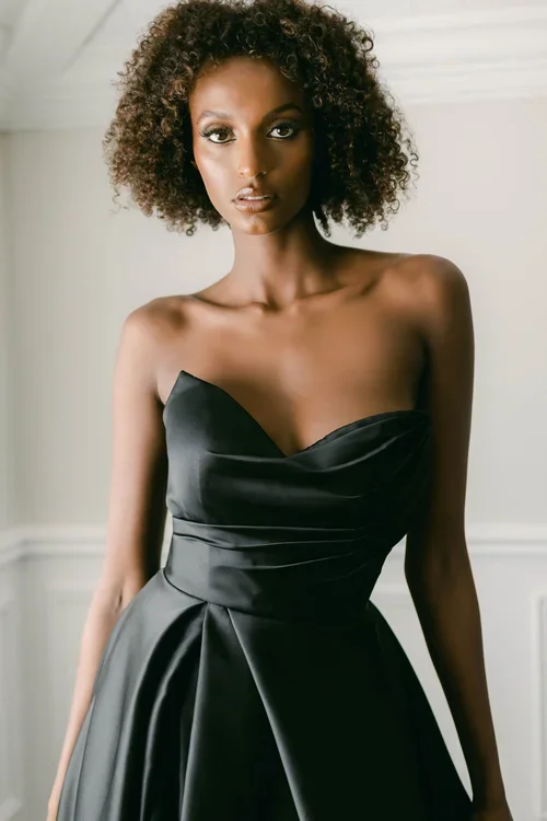 A woman with curly hair wears a strapless, EB22456-1Z black satin gown with a fitted bodice and flared skirt, standing indoors against a light-colored wall.