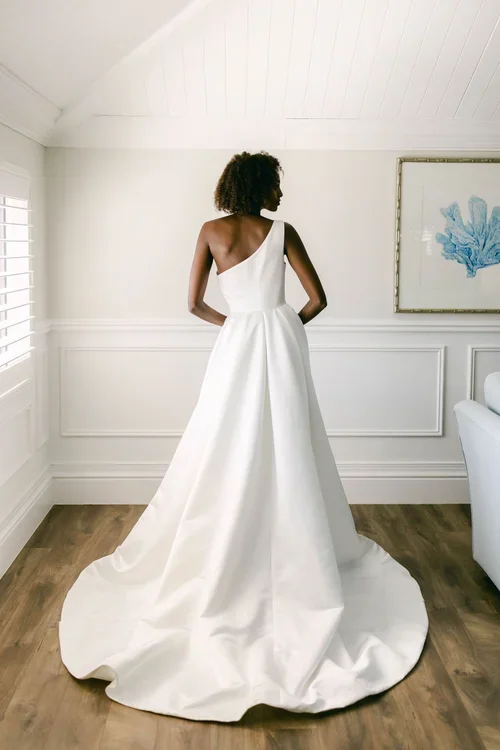 A woman in a white EB22531-1Z one-shoulder bridal gown stands indoors on wooden flooring, facing away from the camera. The dress features a long train, and the room is bright with white walls and a framed artwork.