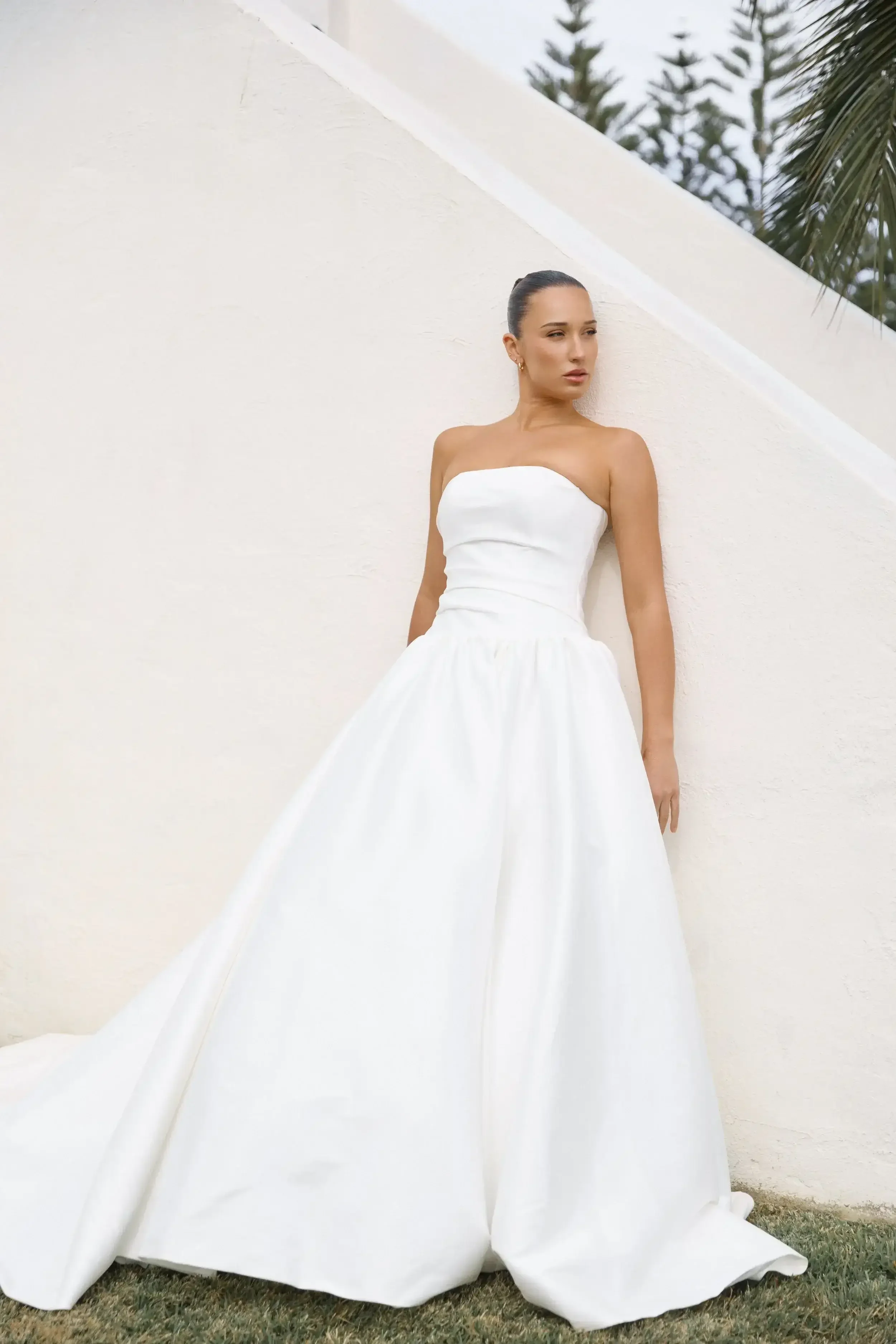 A woman wears the Sur Elle ROSETTE - EB23231 strapless, white, floor-length wedding gown outdoors against a white wall.