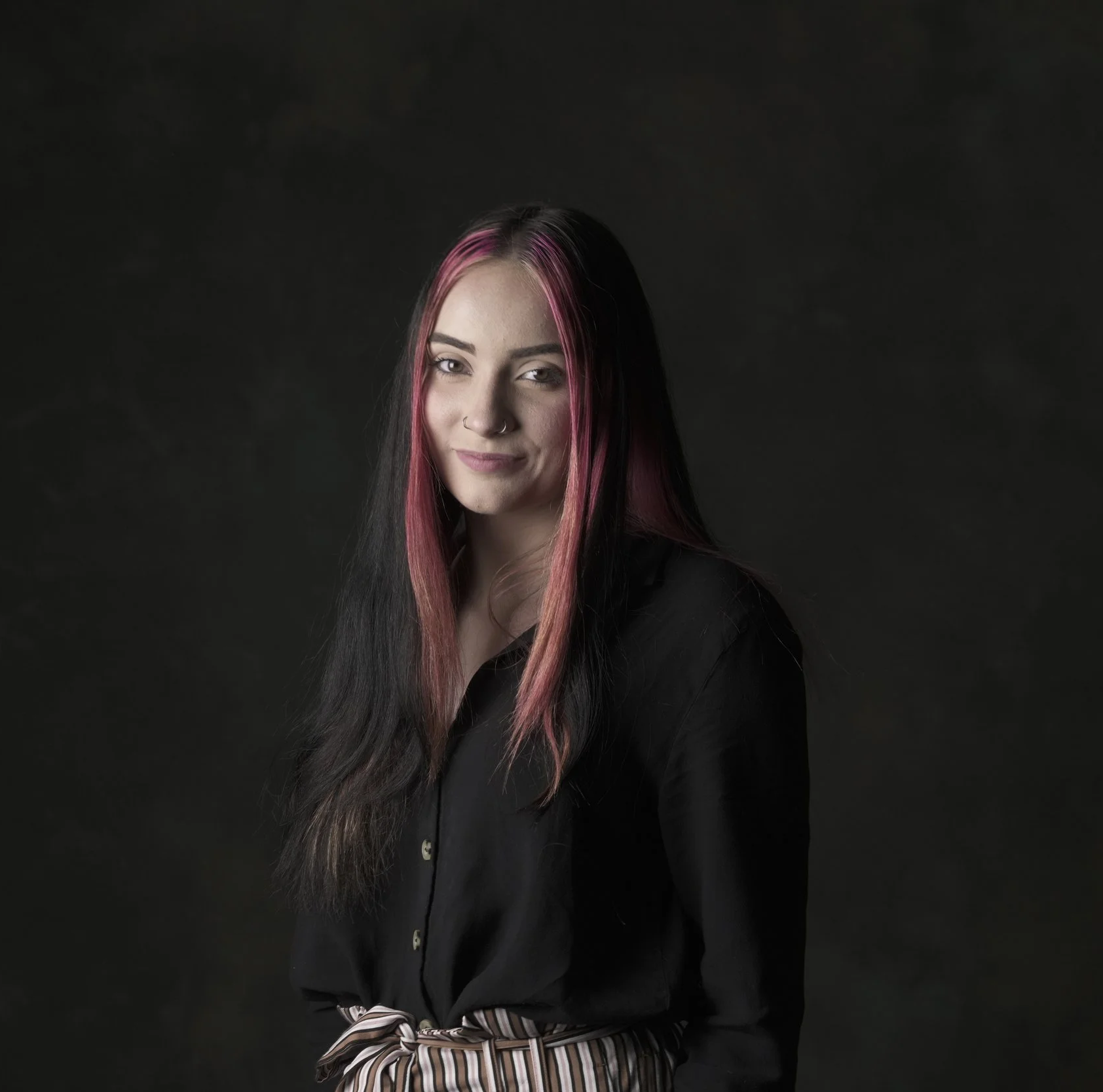 A young woman with long black hair with pink streaks, wearing a black shirt and striped pants, standing against a dark background.