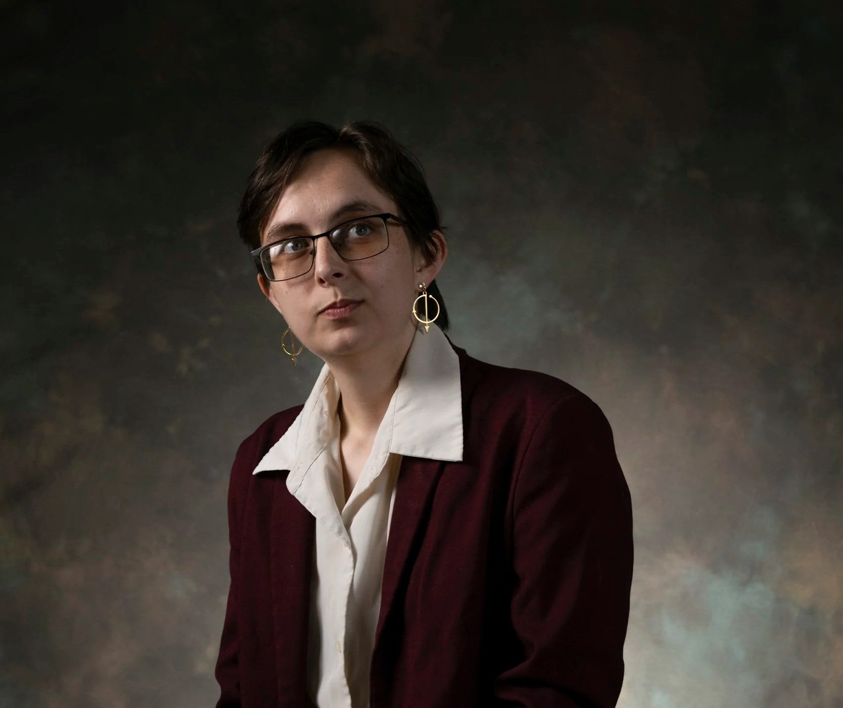 Portrait of a young woman with short dark hair, wearing glasses, gold hoop earrings, a white collared shirt, and a burgundy blazer against a dark, textured background.