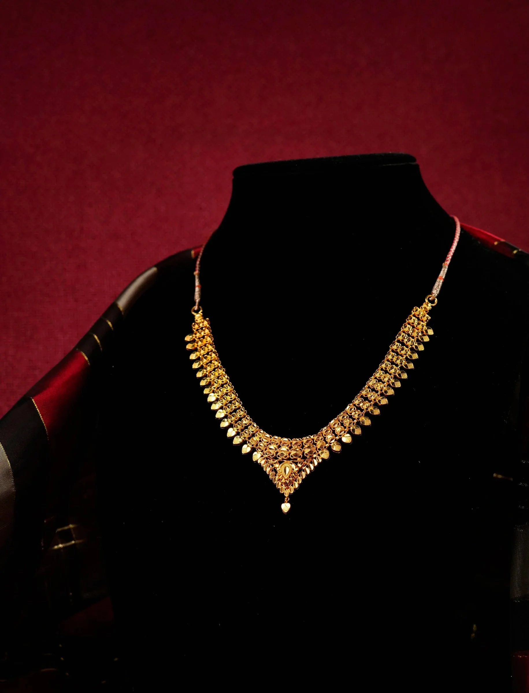 Gold necklace displayed on a black velvet bust against a dark red background.