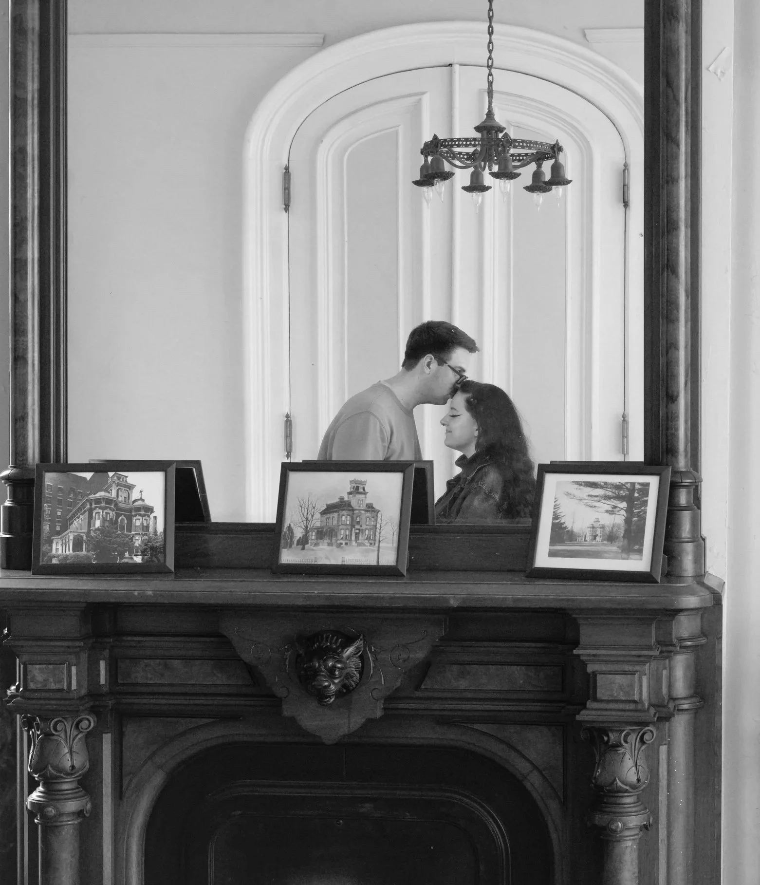 A black and white photo of a mirror above a fireplace, reflecting a couple about to kiss. The mirror has an ornate wooden frame, and three framed pictures of buildings and nature are placed on the fireplace mantel. A chandelier hangs from the ceiling