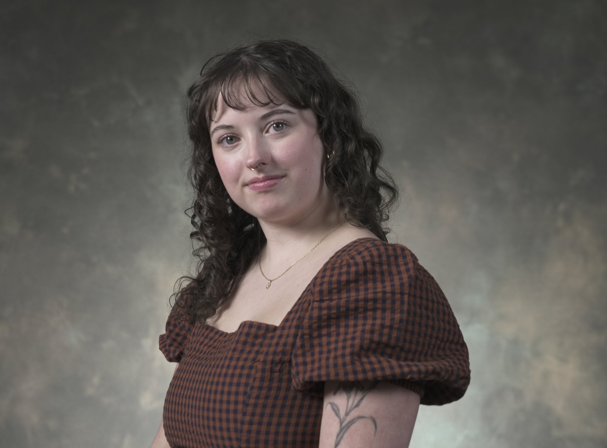 A young woman with wavy dark hair, light skin, and a nose piercing, wearing a brown and black checkered top, standing against a neutral background.