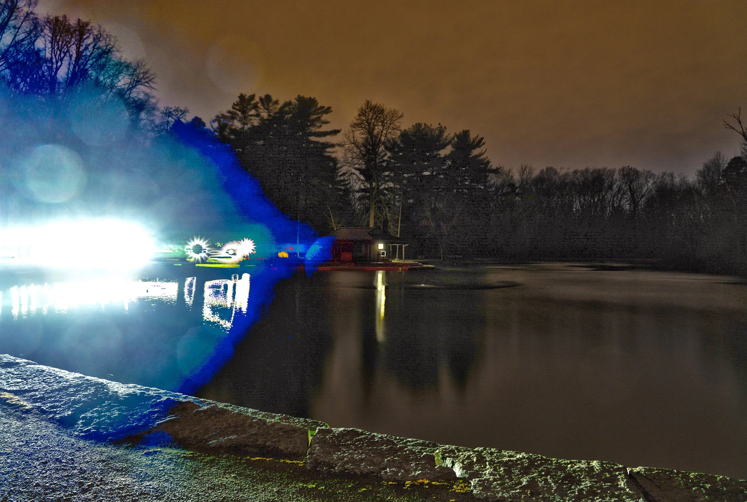 Nighttime scene of a body of water with reflections, a small building with a light, and trees in the background. Bright blue and white light effects are present on the left side of the image.
