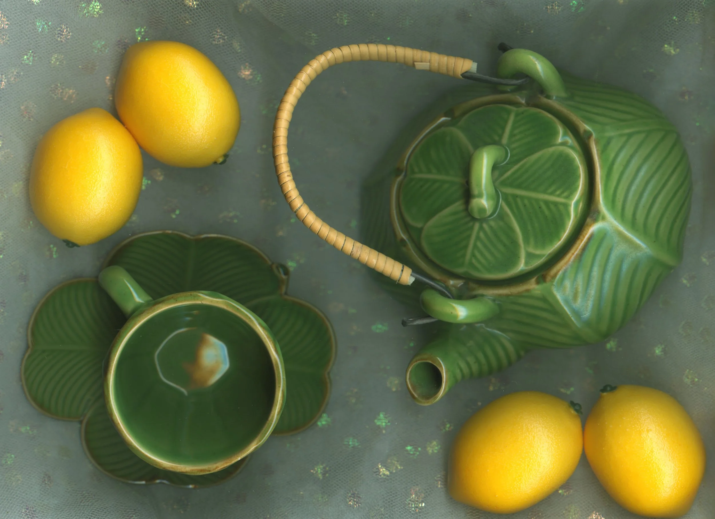 Green and yellow ceramic teapot with a handle, matching teacup, and three yellow lemons arranged on a surface.