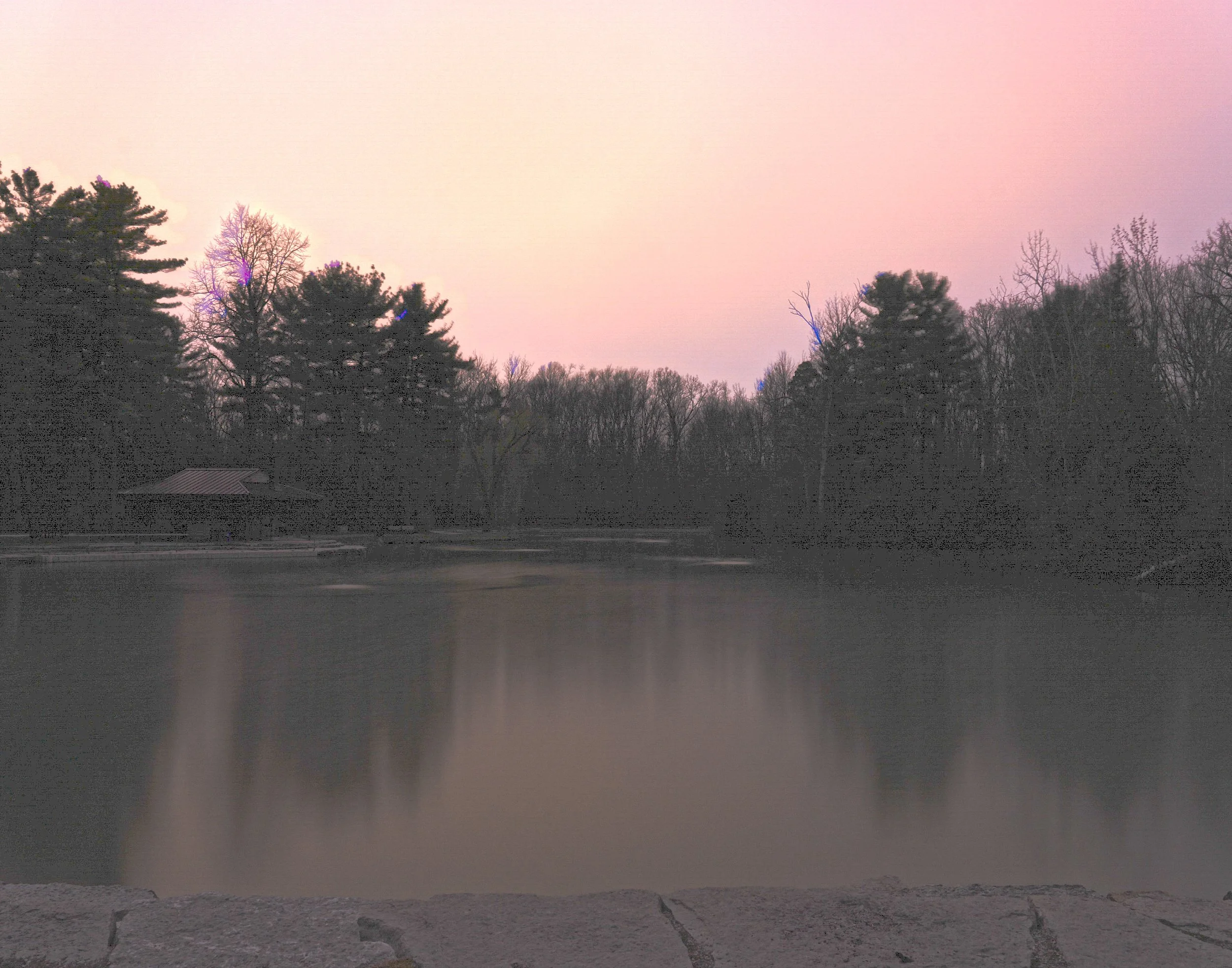 A serene lake scene at dusk with trees surrounding the water and a small pavilion on the left side, pinkish sky reflected in the water.