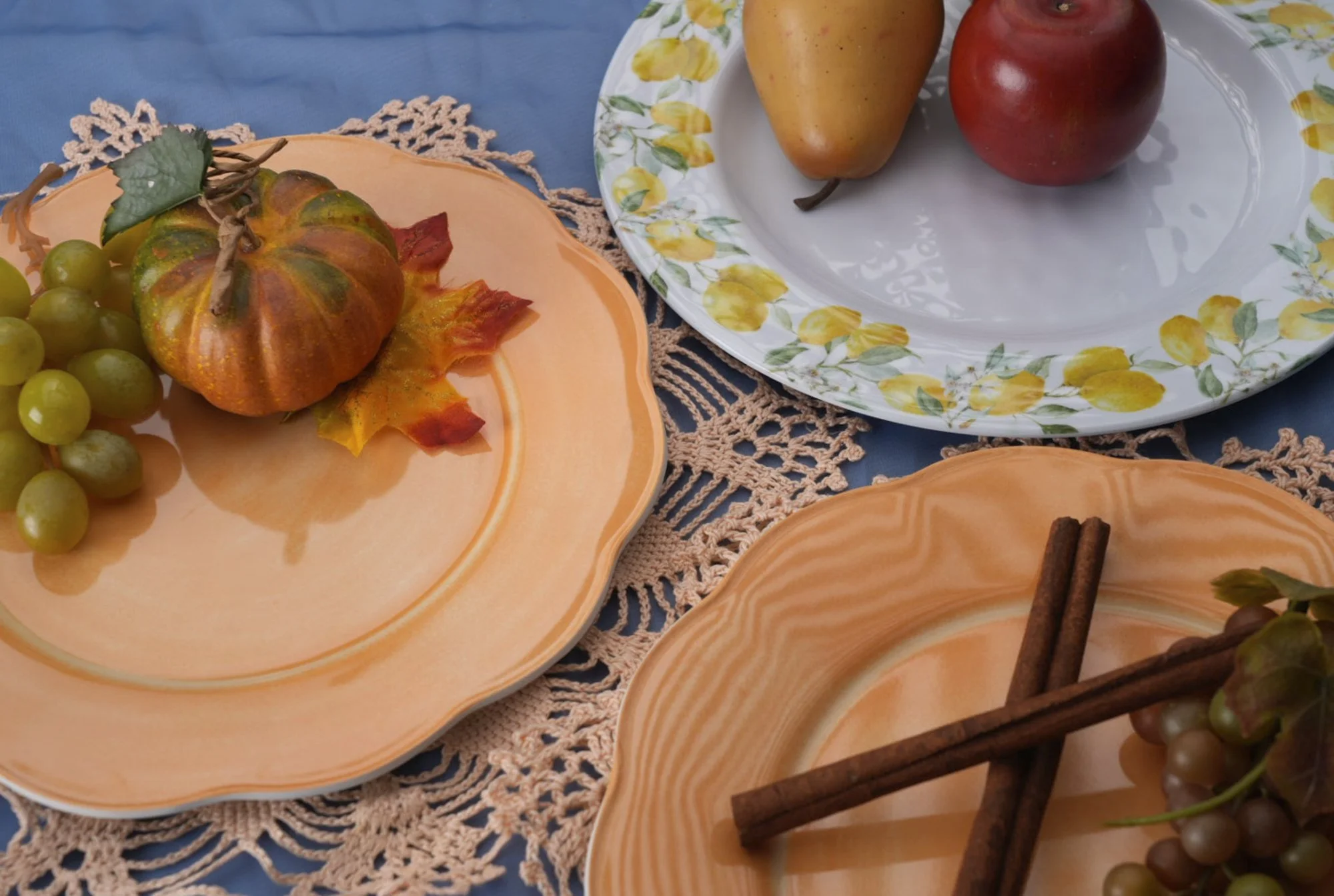 A table with three decorative plates displaying various fruits and spices. The plate on the left holds green grapes, a small ornamental pumpkin, and a decorative maple leaf. The upper right plate contains a pear and an apple. The lower right plate ha