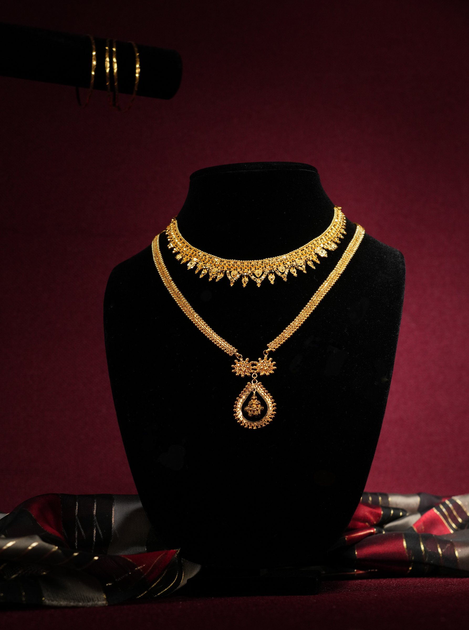 Gold jewelry set displayed on a black velvet bust, including a necklace with intricate design, matching earrings, and a choker, with a red maroon background and a piece of fabric with black, red, and gold accents at the base.