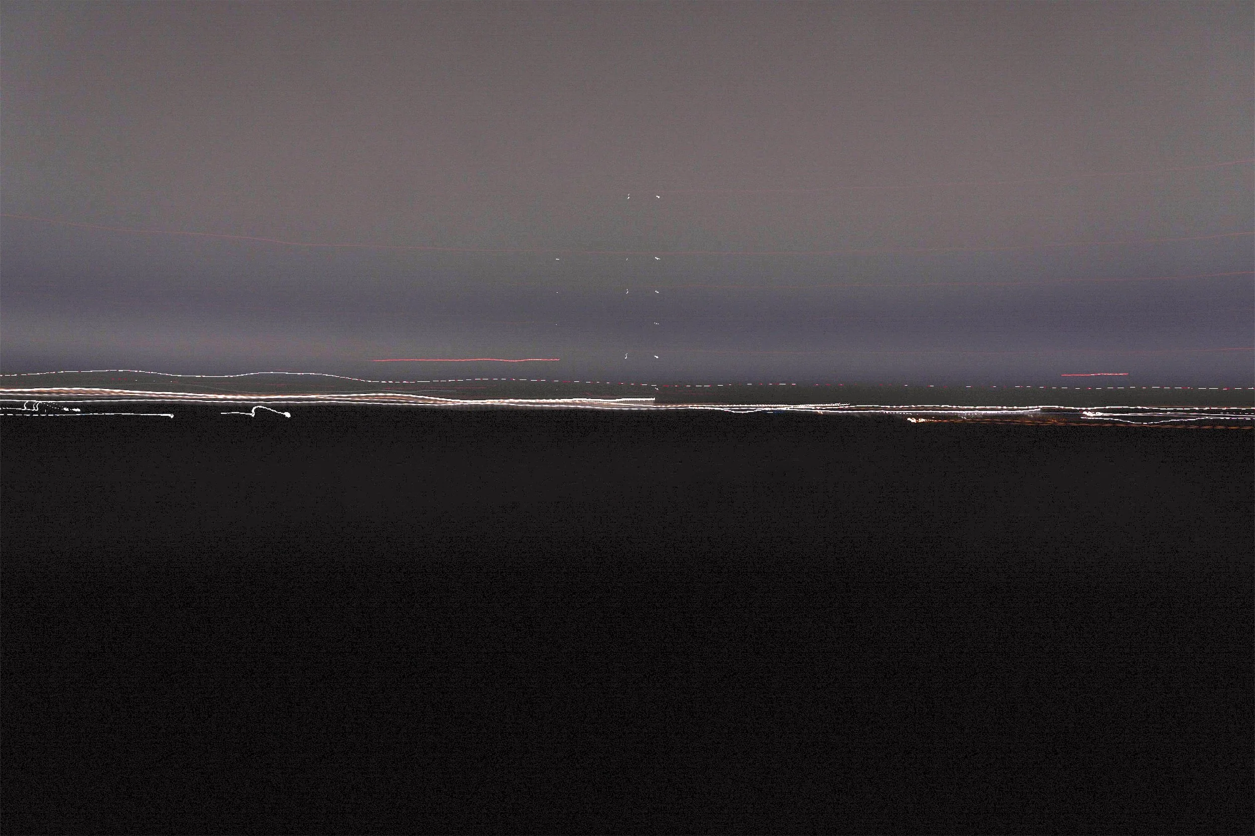 Long exposure night shot of a city skyline with streaks of light from moving vehicles, dark sky with some visible stars, and a dark foreground.