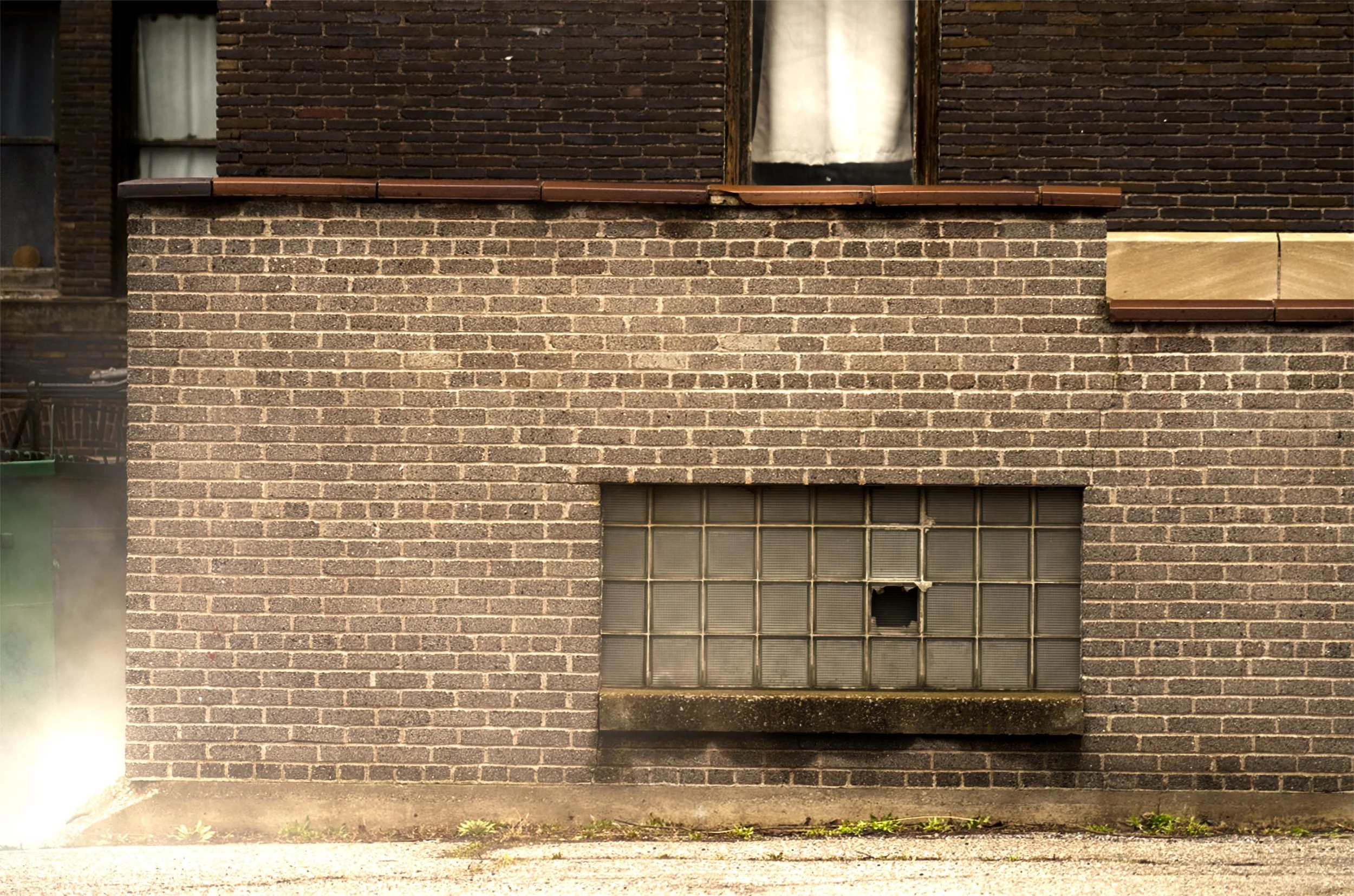 Brick building wall with a broken glass block window and a small hole. The wall has a darker upper section and a lighter lower section, with some greenery and dirt at the base.