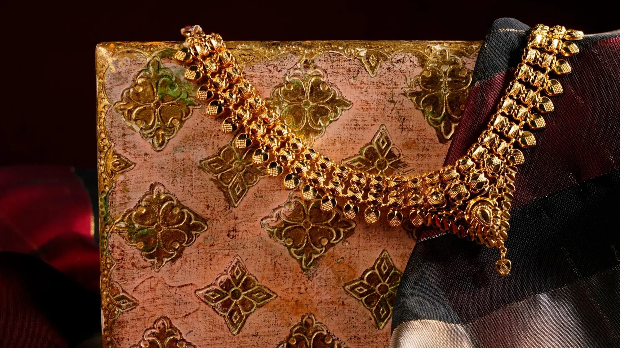 Gold necklace with intricate design draped over a pink and gold patterned gift box, with fabric in black, red, and beige in the background.