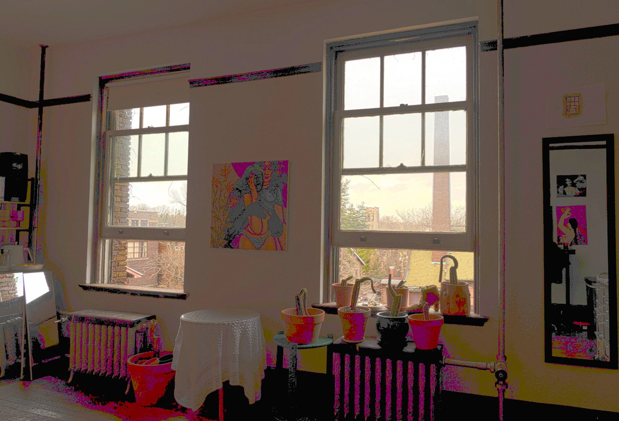 Interior of a room with two large windows, plants in pots on a black radiator, a small white table draped with a cloth, artwork hung on the wall, and framed photos or posters, with a lamp and shelves in the corner.