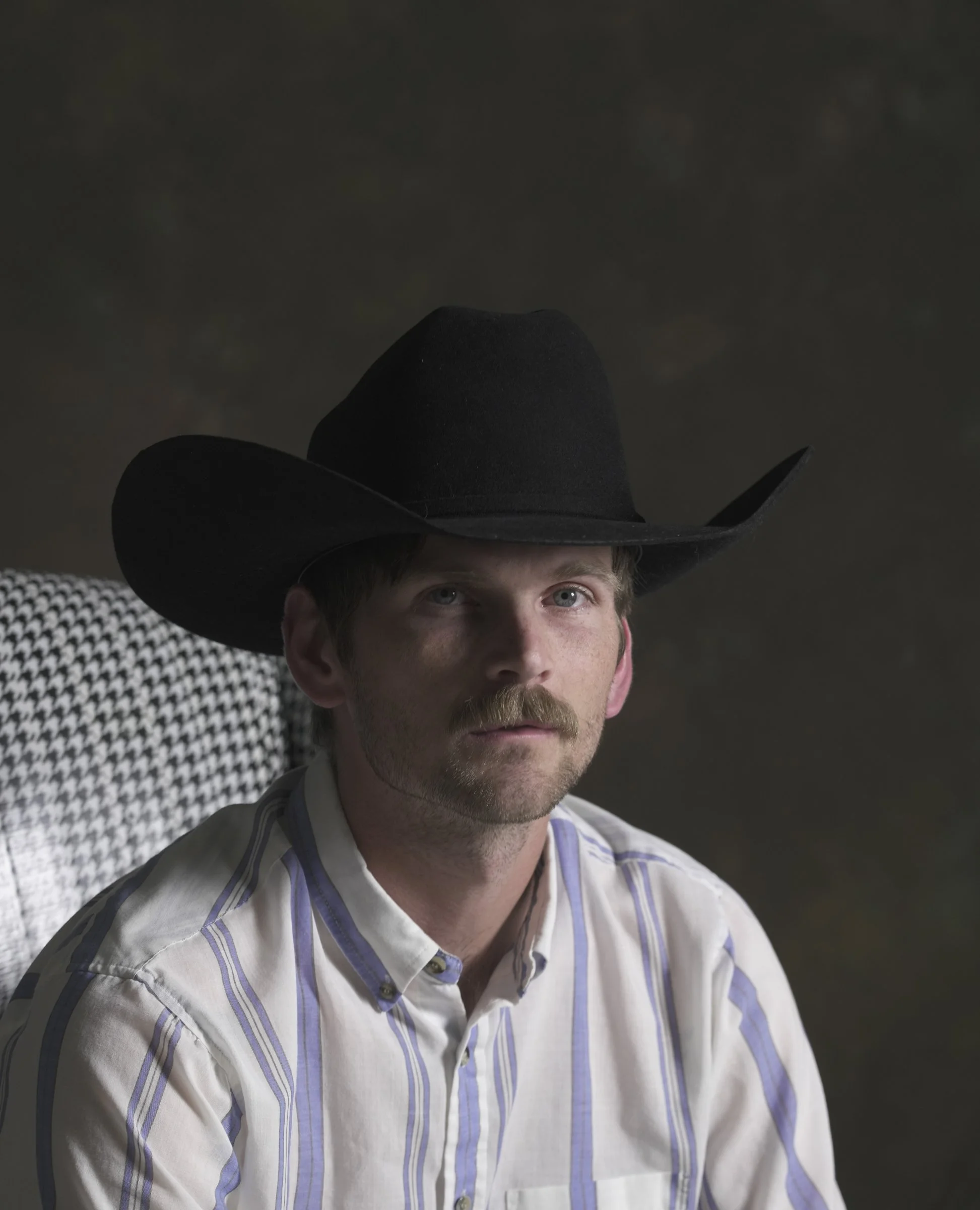 A man wearing a large black cowboy hat, seated against a dark background, with a serious expression, wearing a striped button-up shirt.