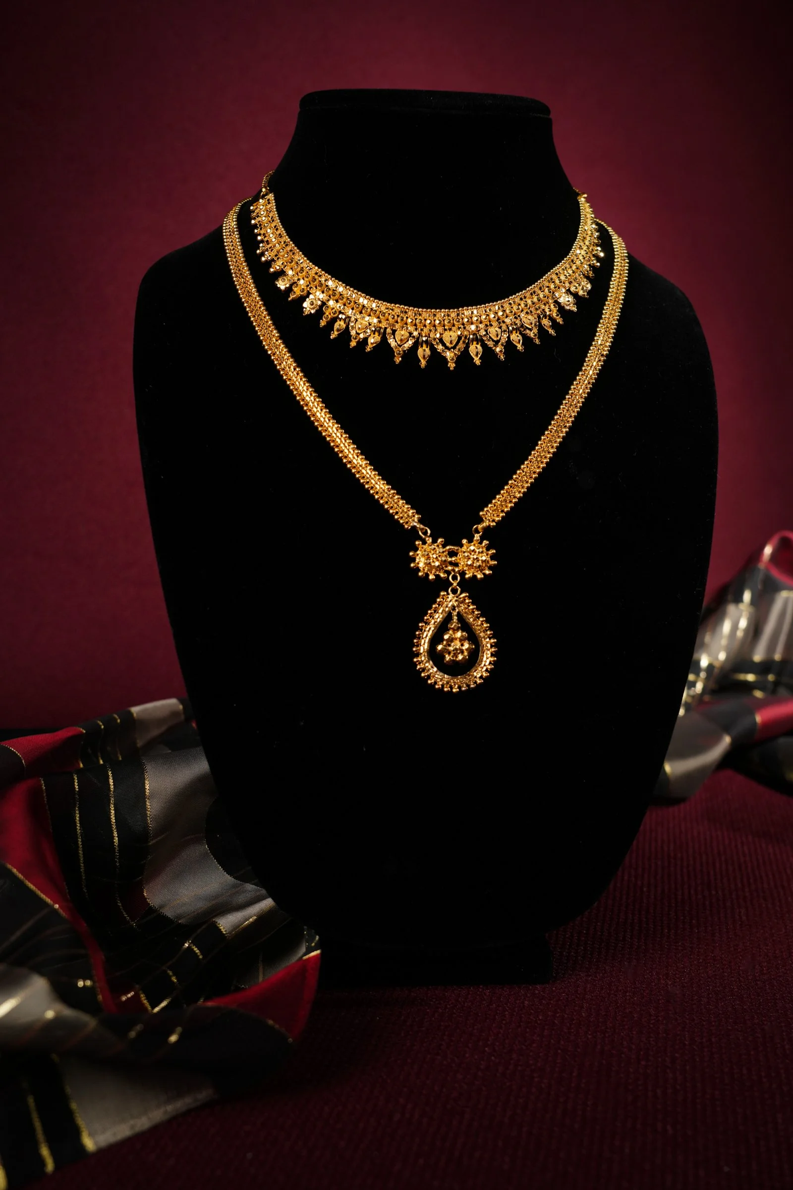 Gold jewelry displayed on a black velvet bust, including layered necklaces and earrings, with patterned fabric beneath.