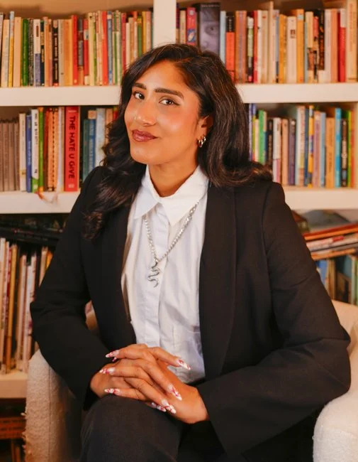 A woman in a black blazer and white shirt sitting in front of a bookshelf filled with books.