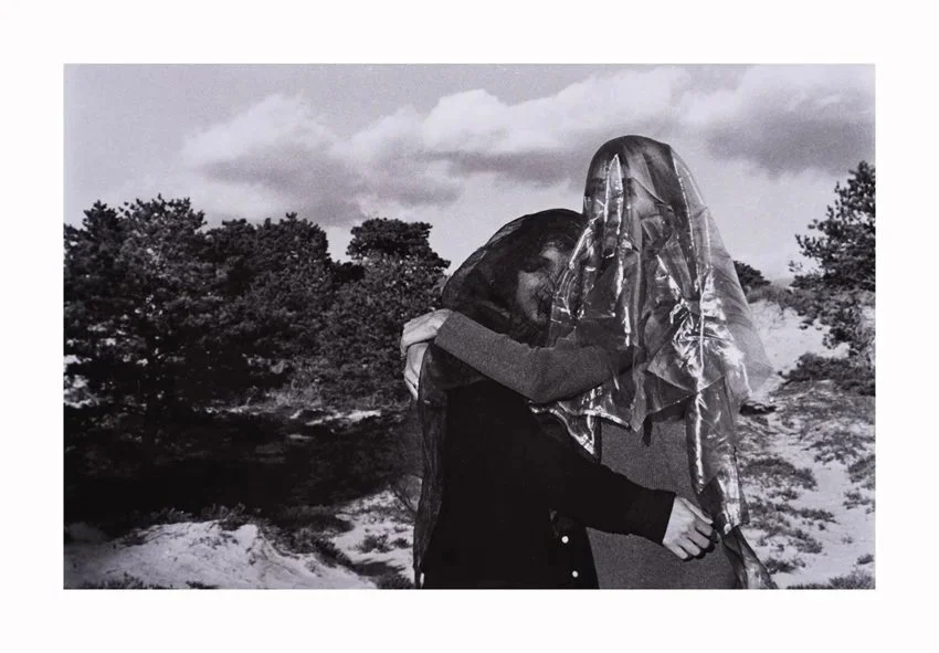 Two people hugging outdoors, one person wearing a metallic plastic bag over their head in a natural landscape with trees and cloudy sky.
