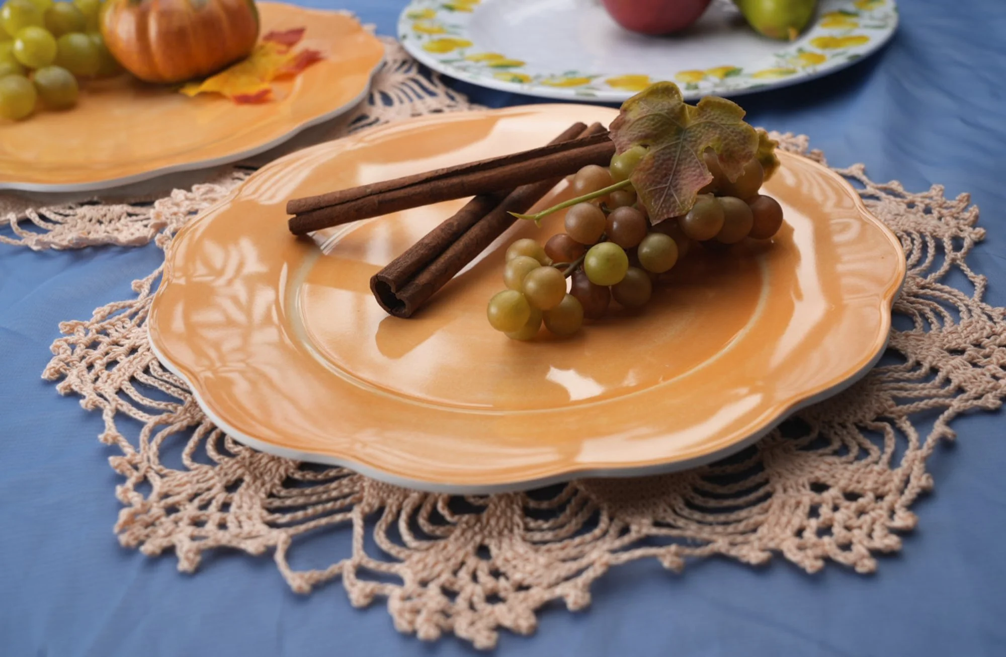 A plate with grapes and cinnamon sticks, on a lace doily and a blue tablecloth, with part of another similar plate in the background.