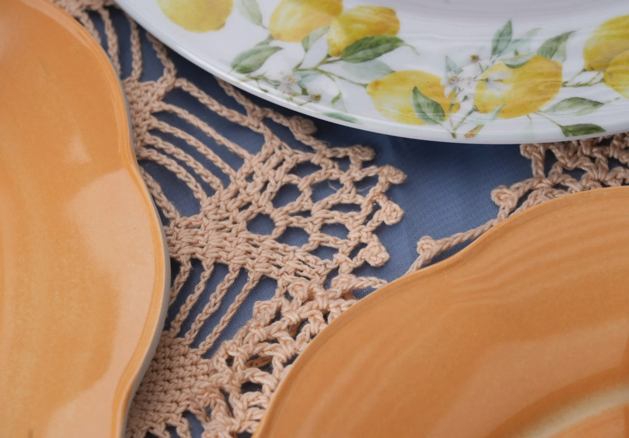 Close-up of a table setting with orange plates, a floral plate with yellow lemons and green leaves, a crocheted tablecloth, and a blue table surface.