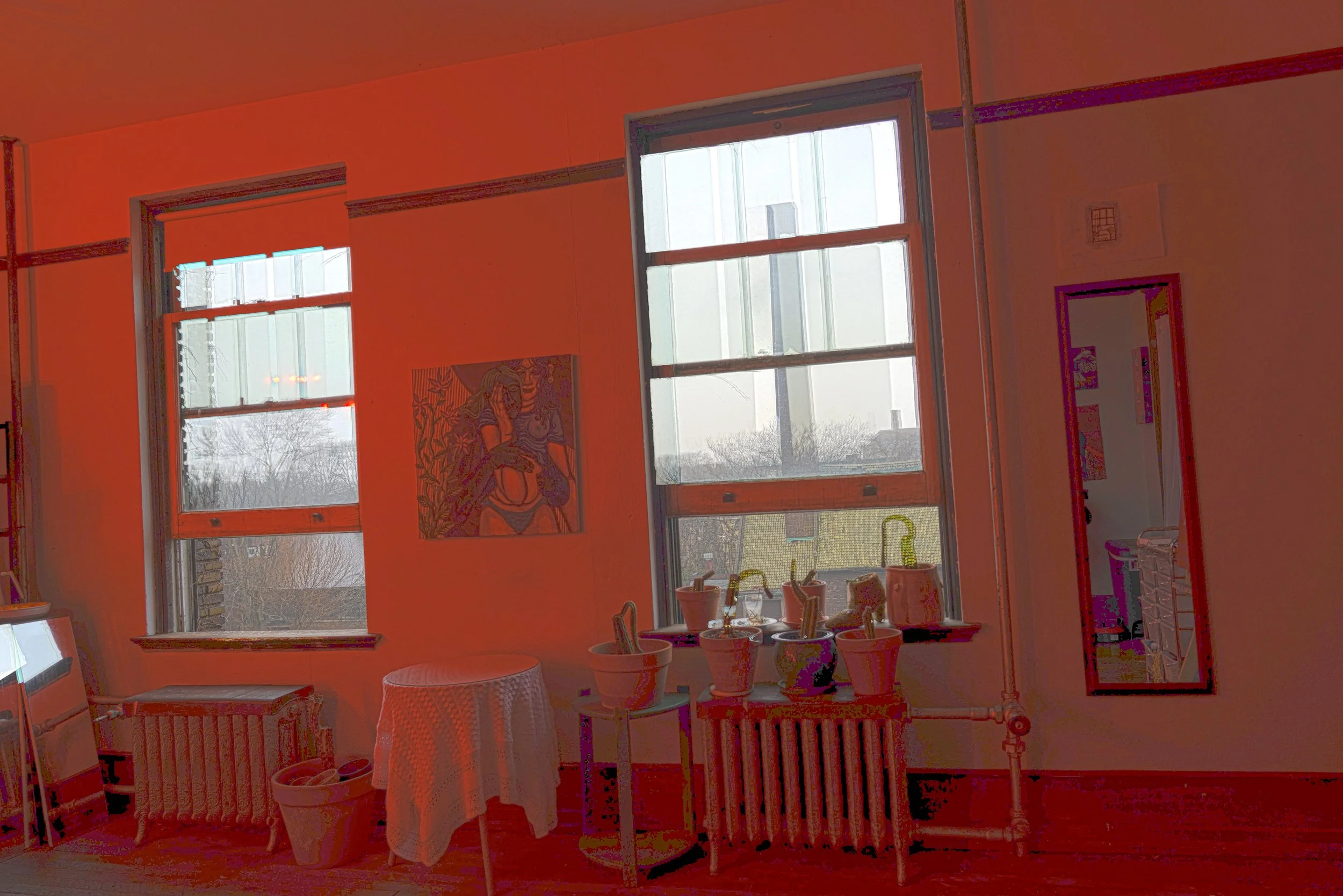 An interior room with two large windows, a colorful painting on the wall, and a mirror on the right. There is a small table with a white cloth, a radiator, and various potted plants on the window sill.