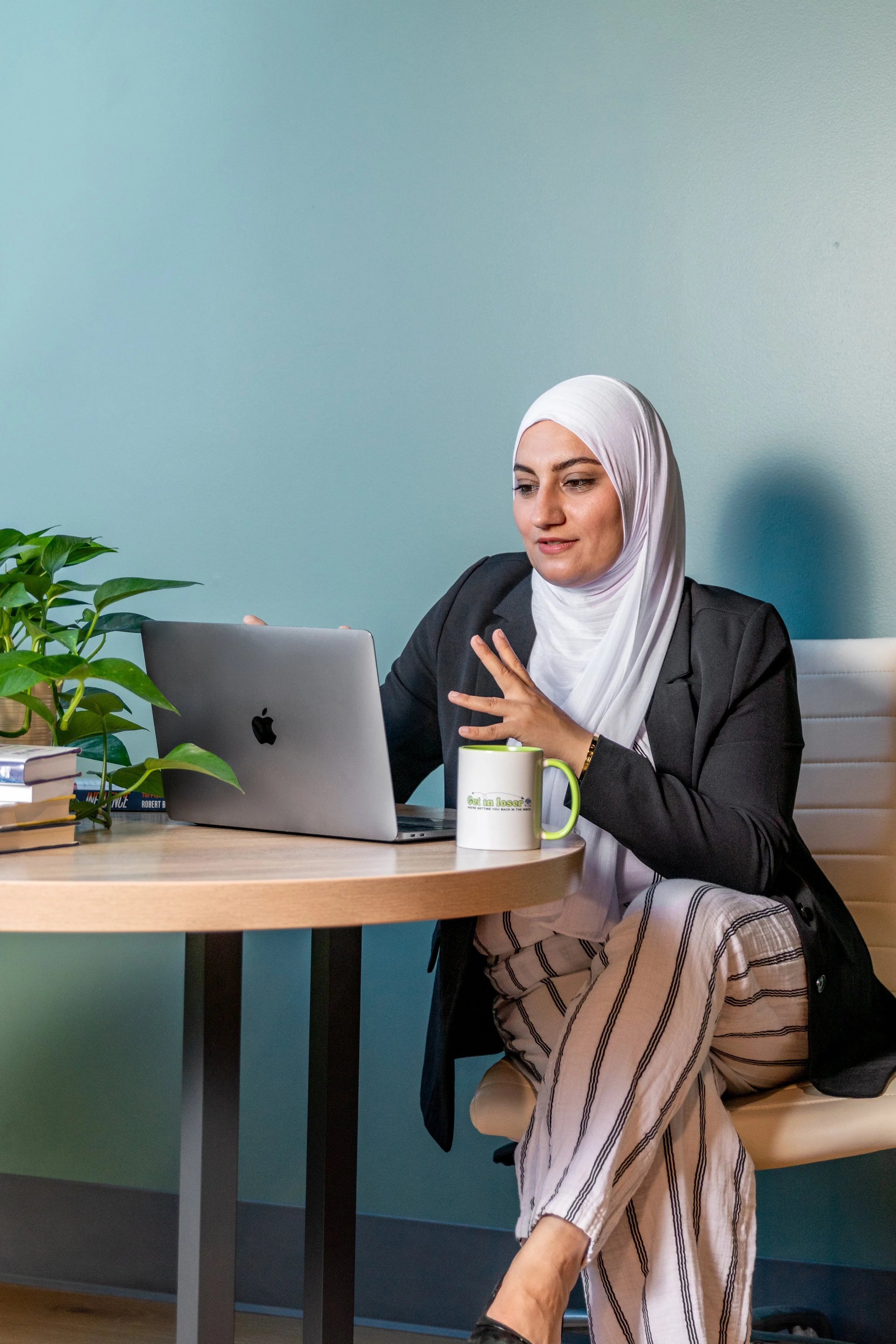 A woman wearing a white headscarf and black blazer sitting at a wooden desk with a silver laptop, green and white coffee mug, and a stack of books, working in an office with a light blue wall.