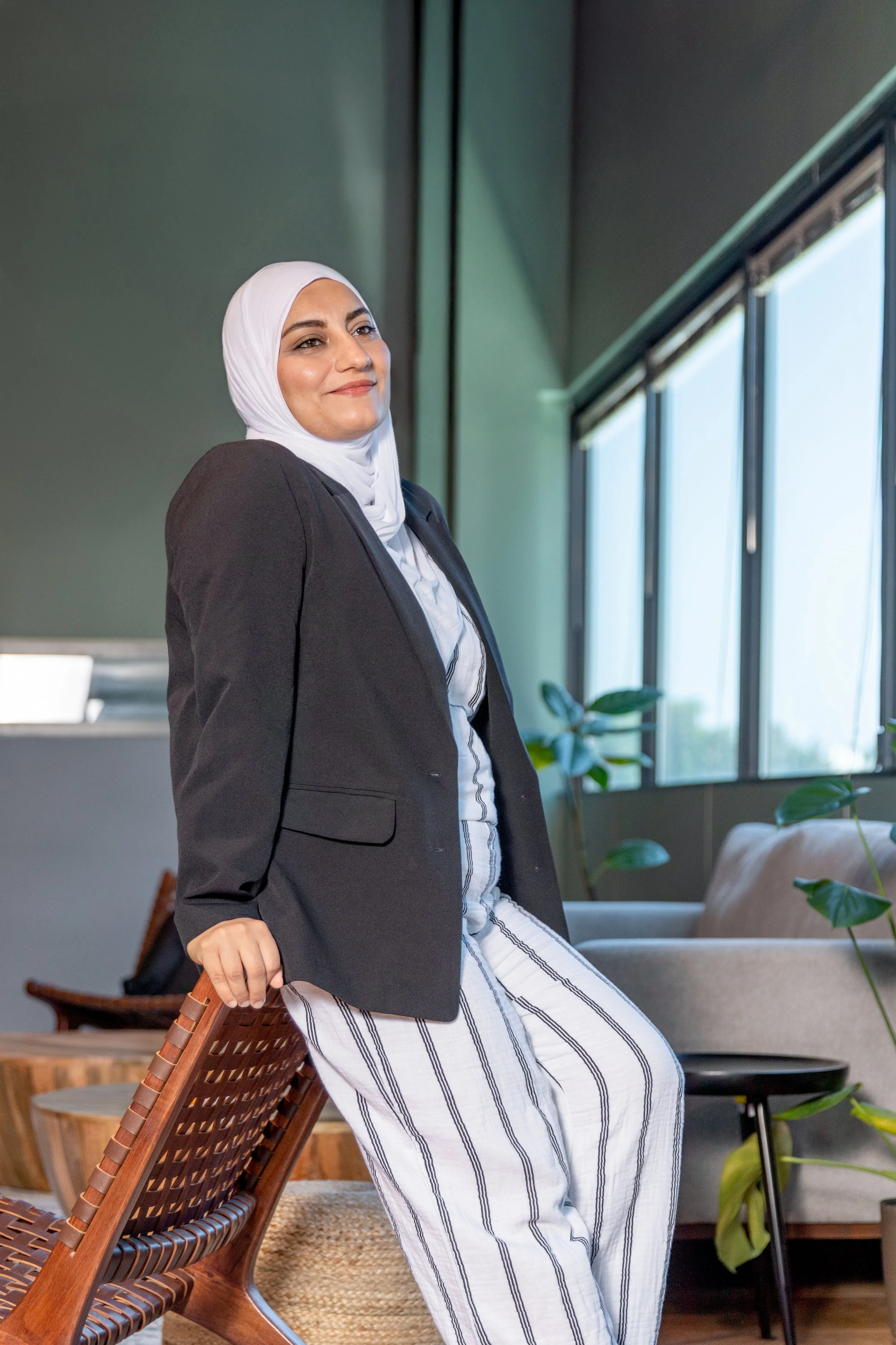 A woman wearing a white hijab, dark blazer, and striped pants is leaning on a wooden chair in a modern, brightly lit room with large windows and green plants.
