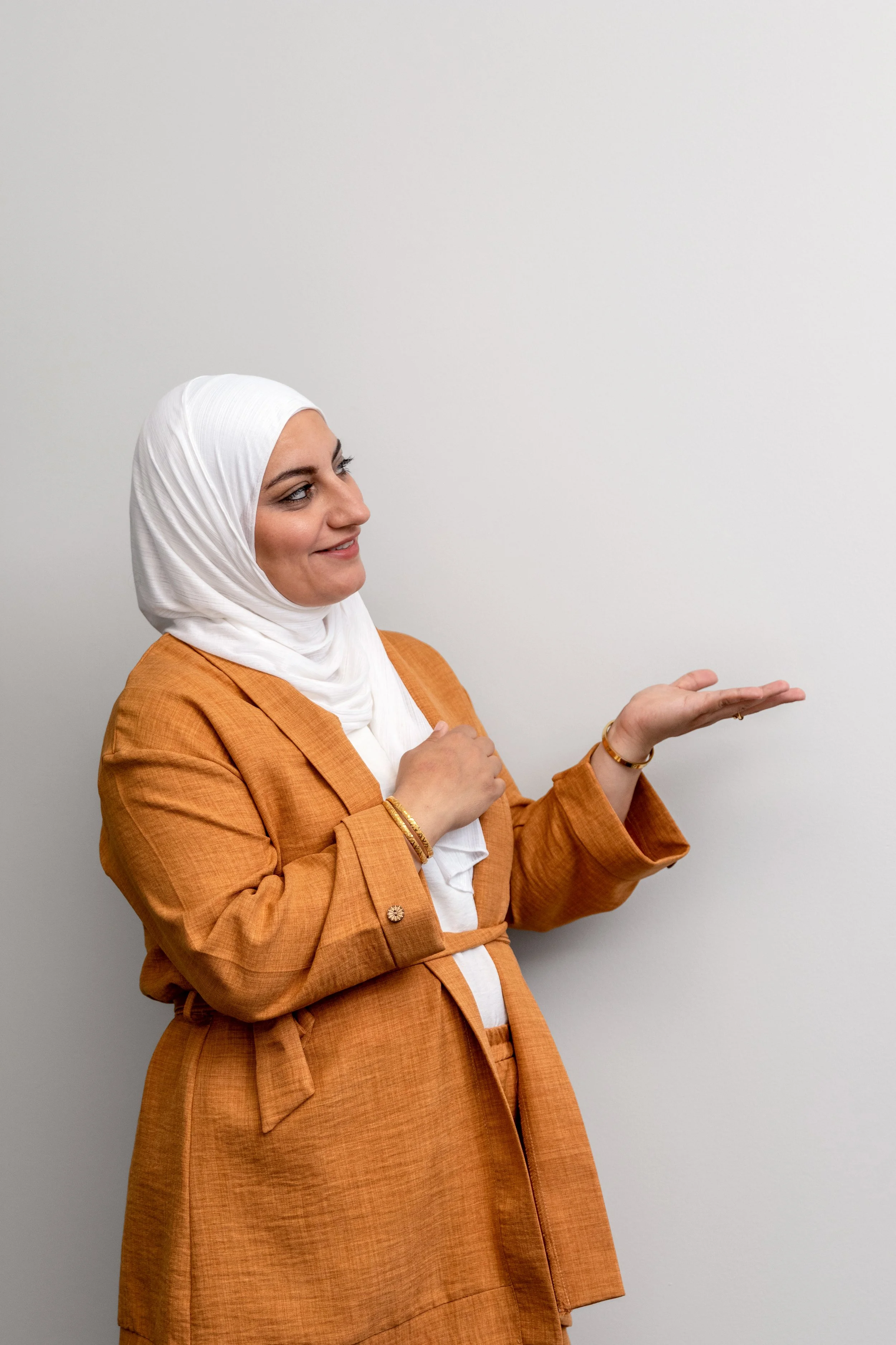 A woman wearing a white hijab and an orange blazer, gesturing with her right hand while smiling and looking to her left, standing against a plain white wall.