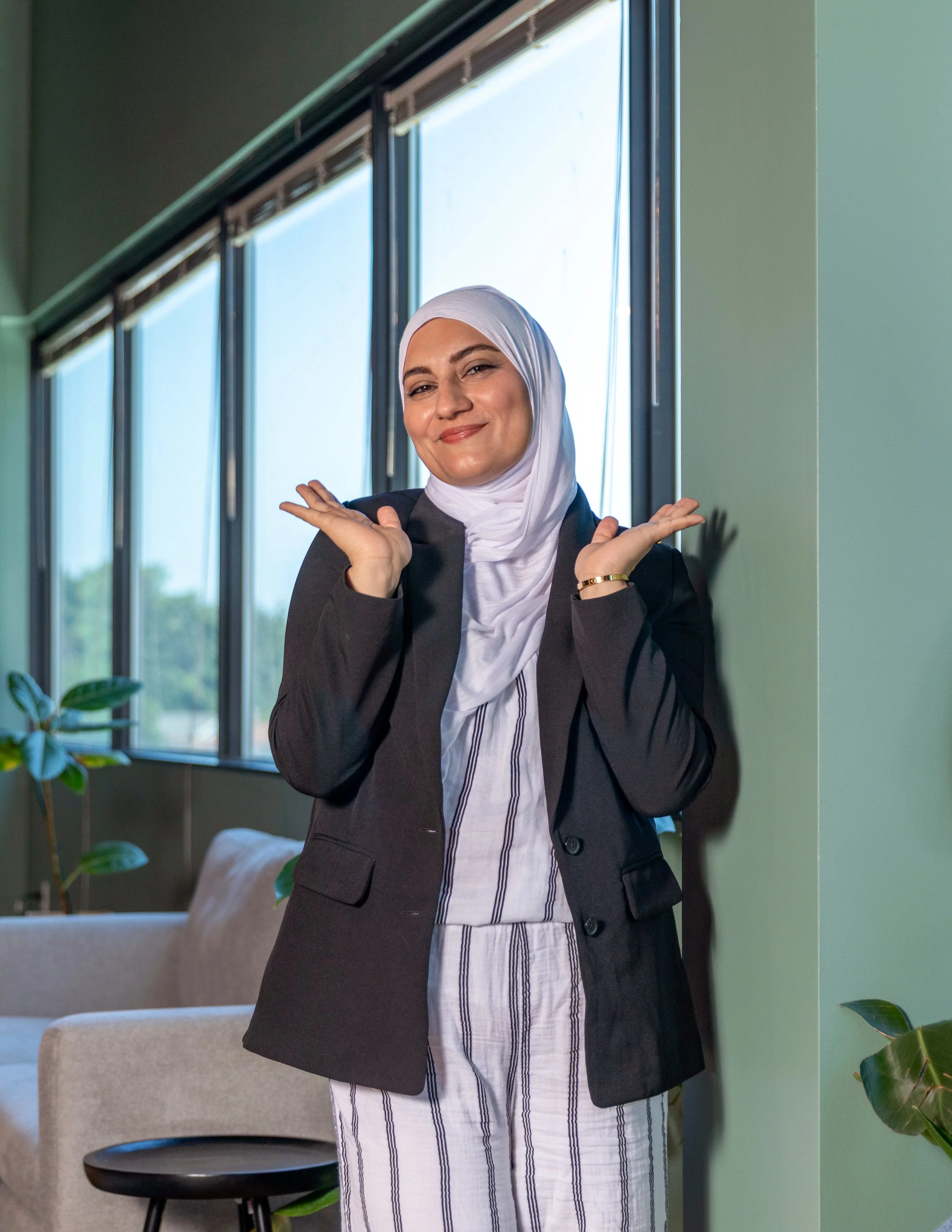A woman wearing a white hijab and a dark blazer standing indoors next to a large window, smiling with hands raised near her face.