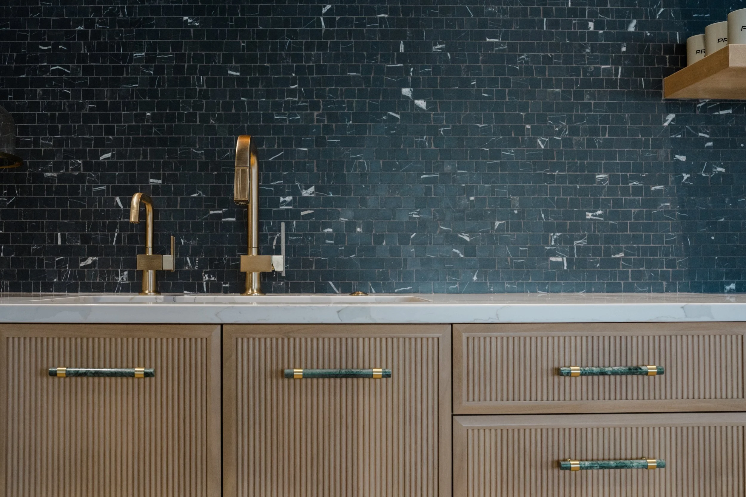 Modern kitchen sink with gold fixtures and black marble tile backsplash, wooden cabinetry, and floating shelf with white containers.