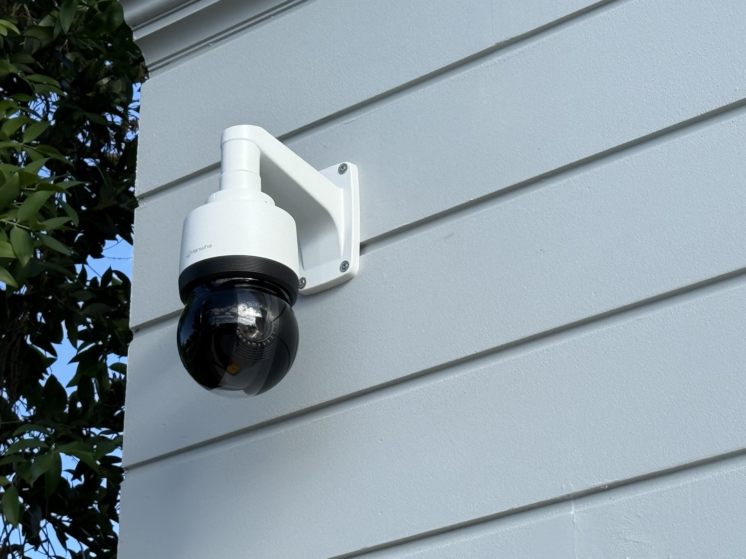 Outdoor security camera mounted on a white wall with nearby foliage.