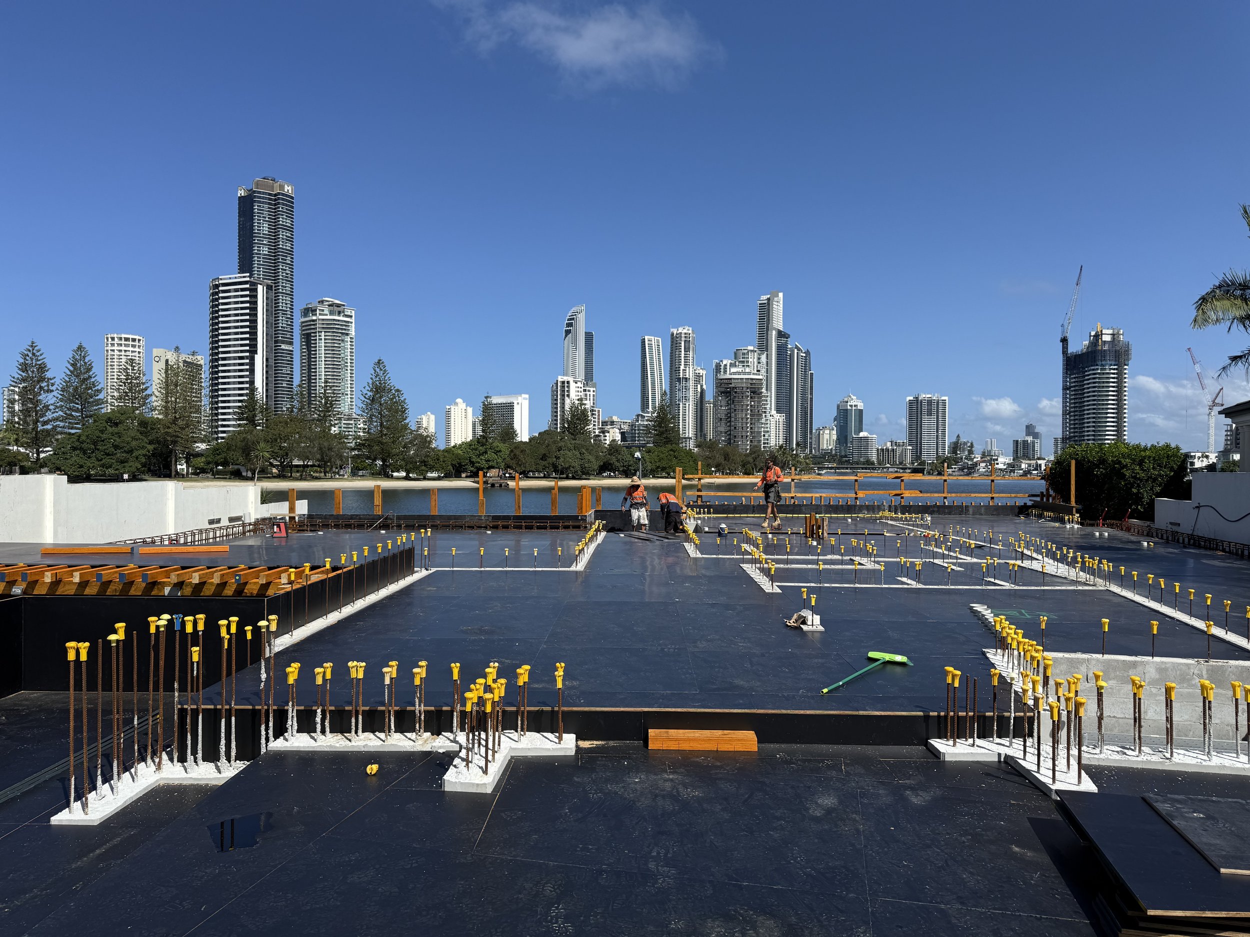 Construction site with unfinished concrete foundation, rebar, and skyscrapers in the background, under a clear blue sky.