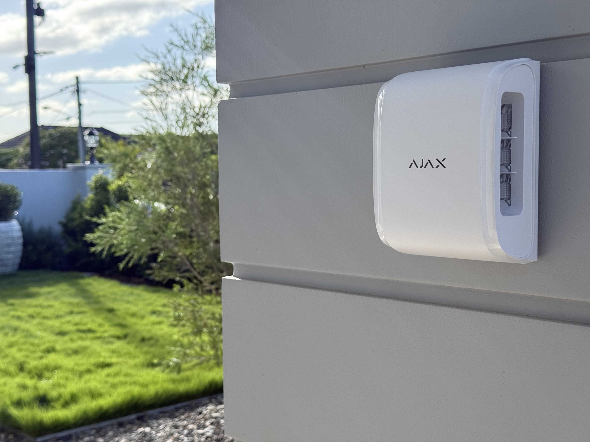 Outdoor security sensor attached to a wall with a garden in the background.