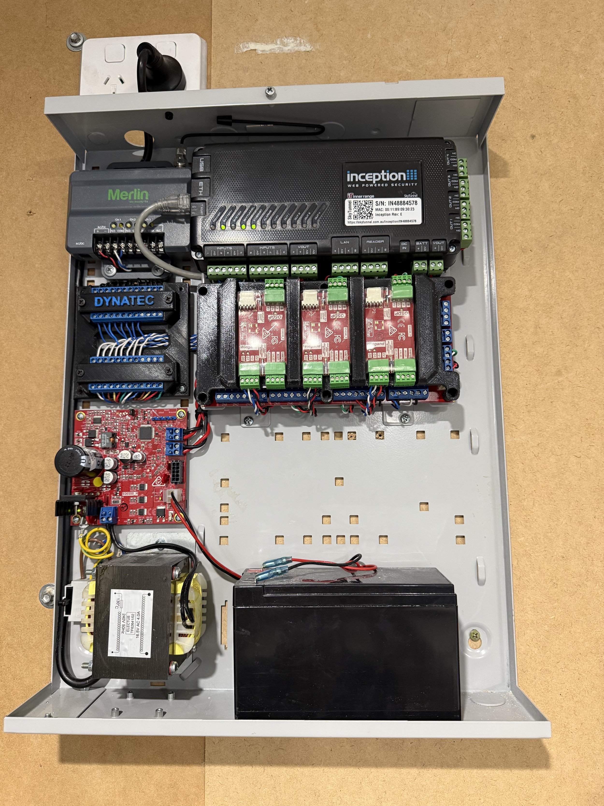 Open security control panel with electronic components and wiring, including a circuit board and power supply, mounted on a wooden surface.