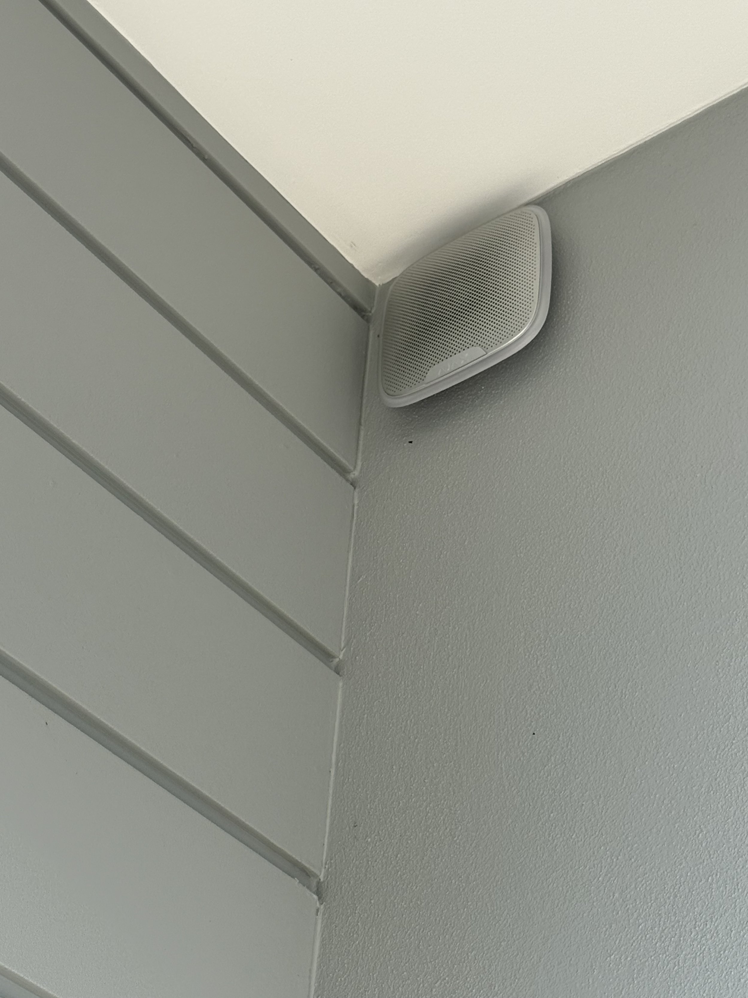 Ceiling-mounted white speaker in corner of a room with gray walls and vertical paneling.