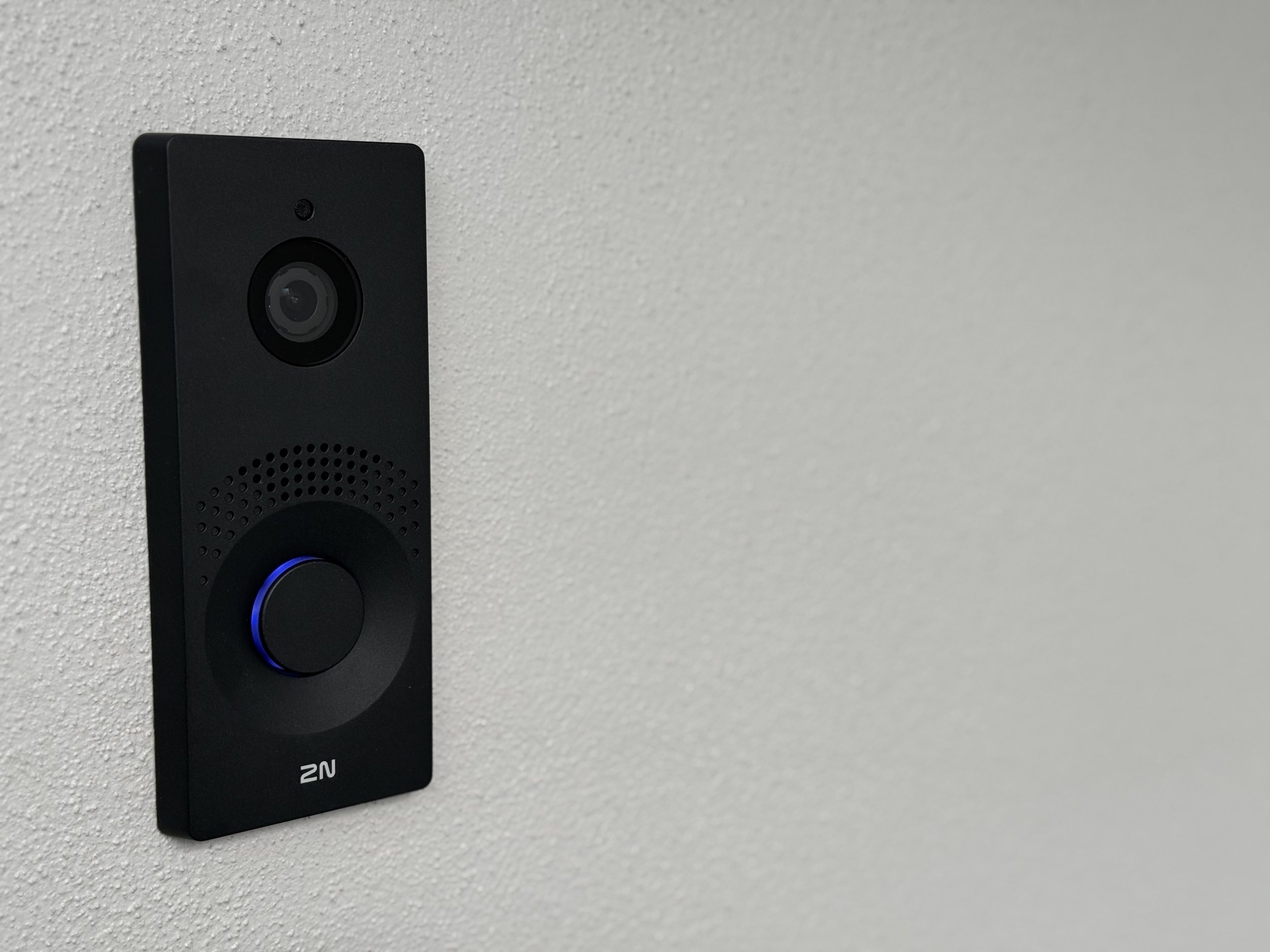 Black video doorbell with a camera and speaker, mounted on a white wall.