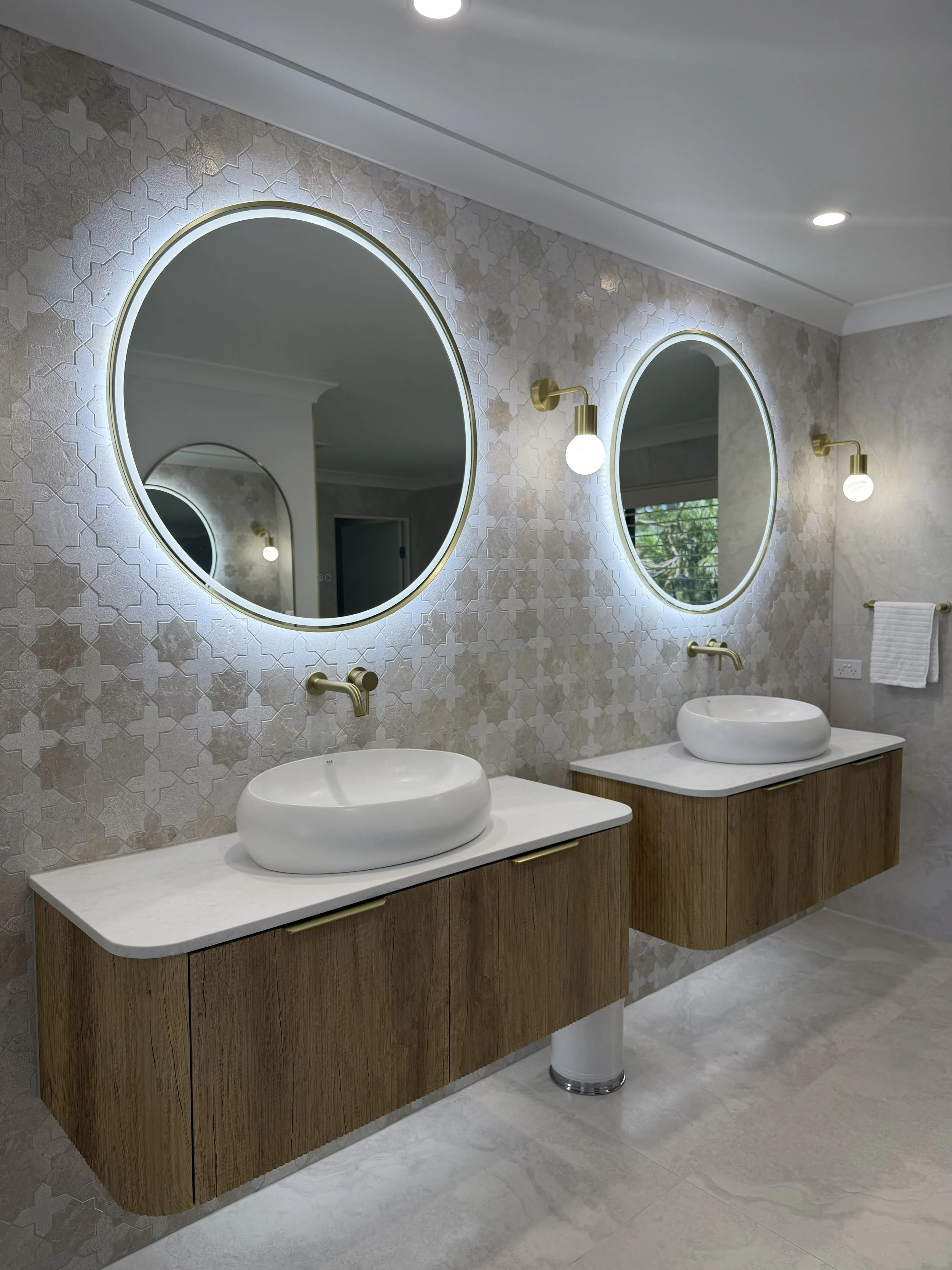 Modern bathroom with two round backlit mirrors, two vessel sinks, gold wall sconces, a patterned wall, and a floating wooden vanity.
