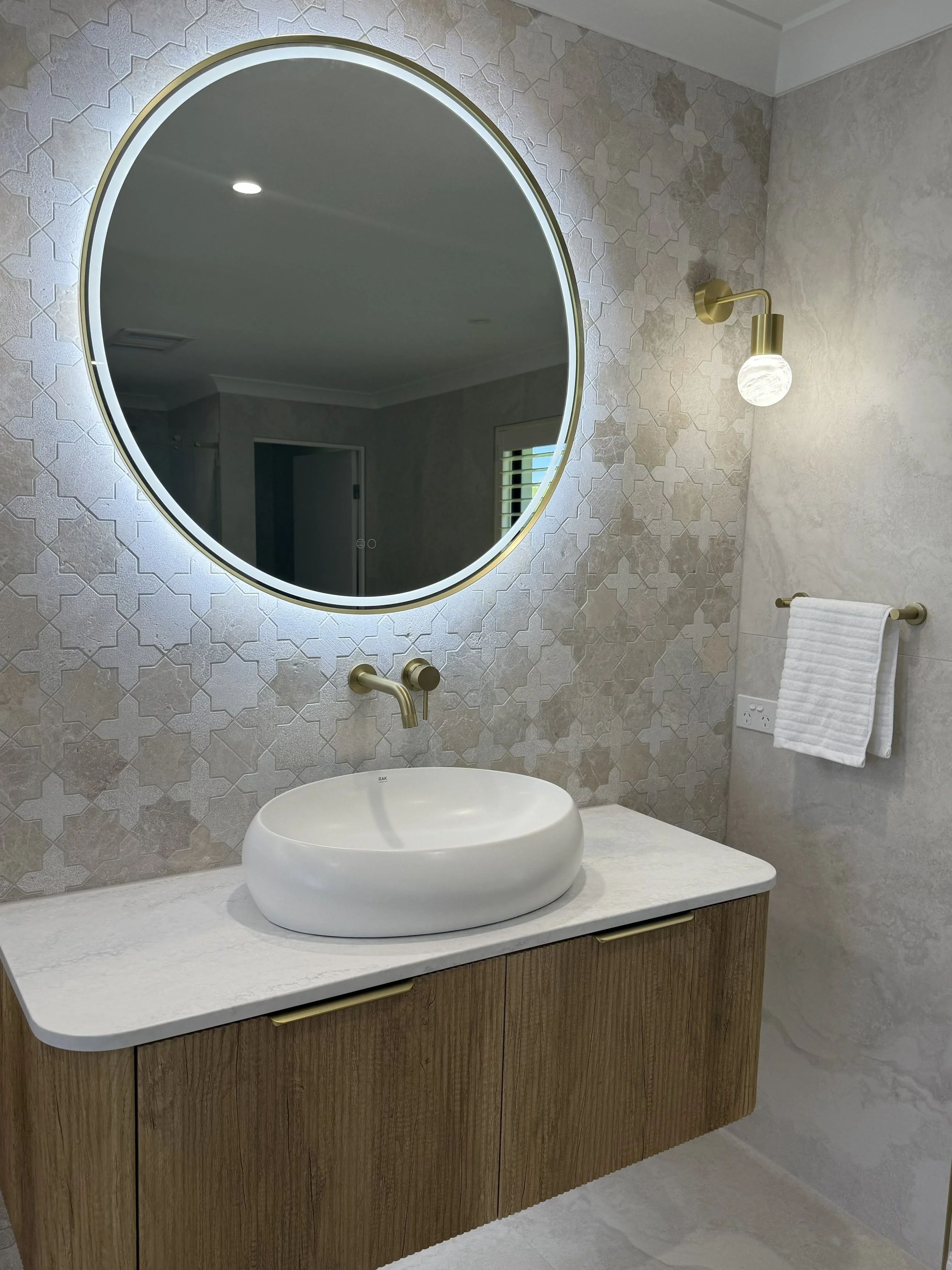 Modern bathroom with a round illuminated mirror, vessel sink, wall-mounted brass faucet, and a wooden vanity. There is a brass wall sconce and a towel hanging on a brass towel bar.