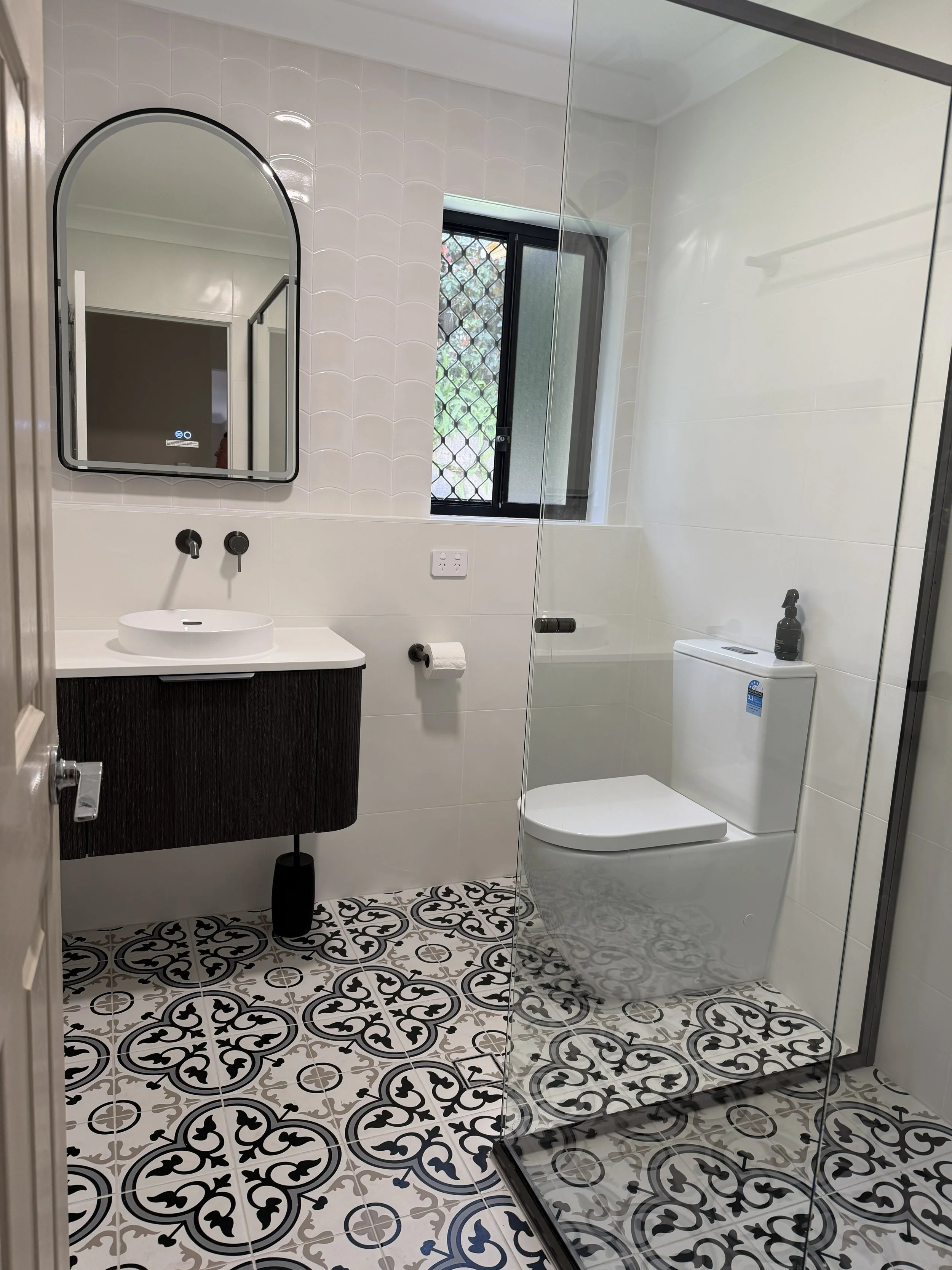 Modern bathroom with patterned black and white floor tiles, a white toilet, a black vanity with a vessel sink, a mirror, a window with security bars, and a glass-enclosed shower.