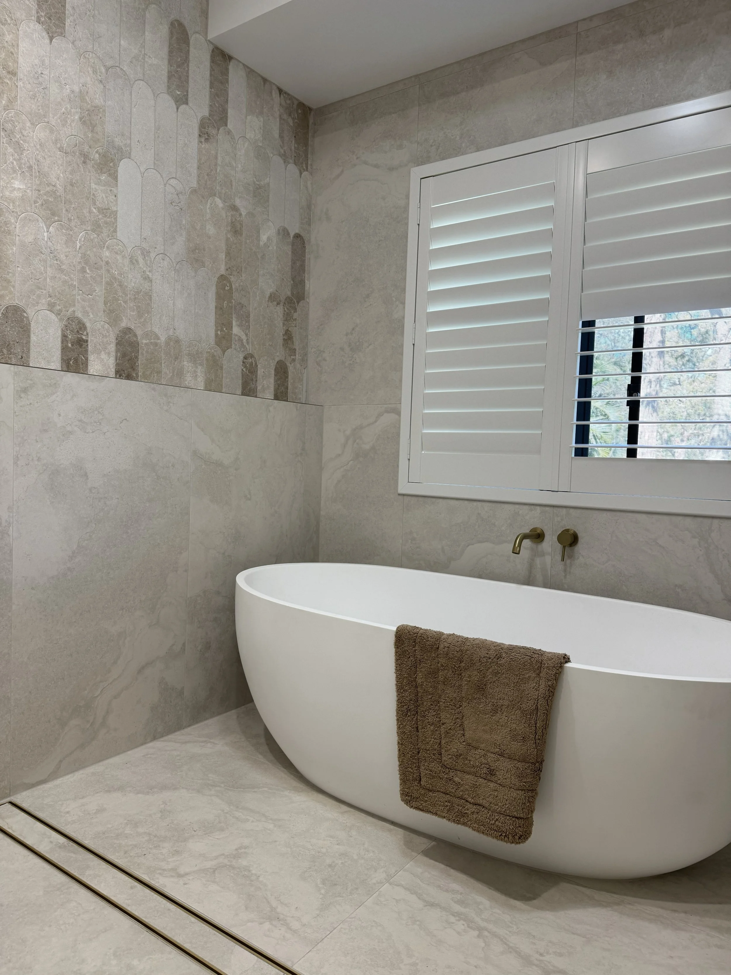 A modern bathroom with a freestanding white bathtub positioned under a window with white plantation shutters, beige and gray tiled walls, and a brown towel hanging over the tub.