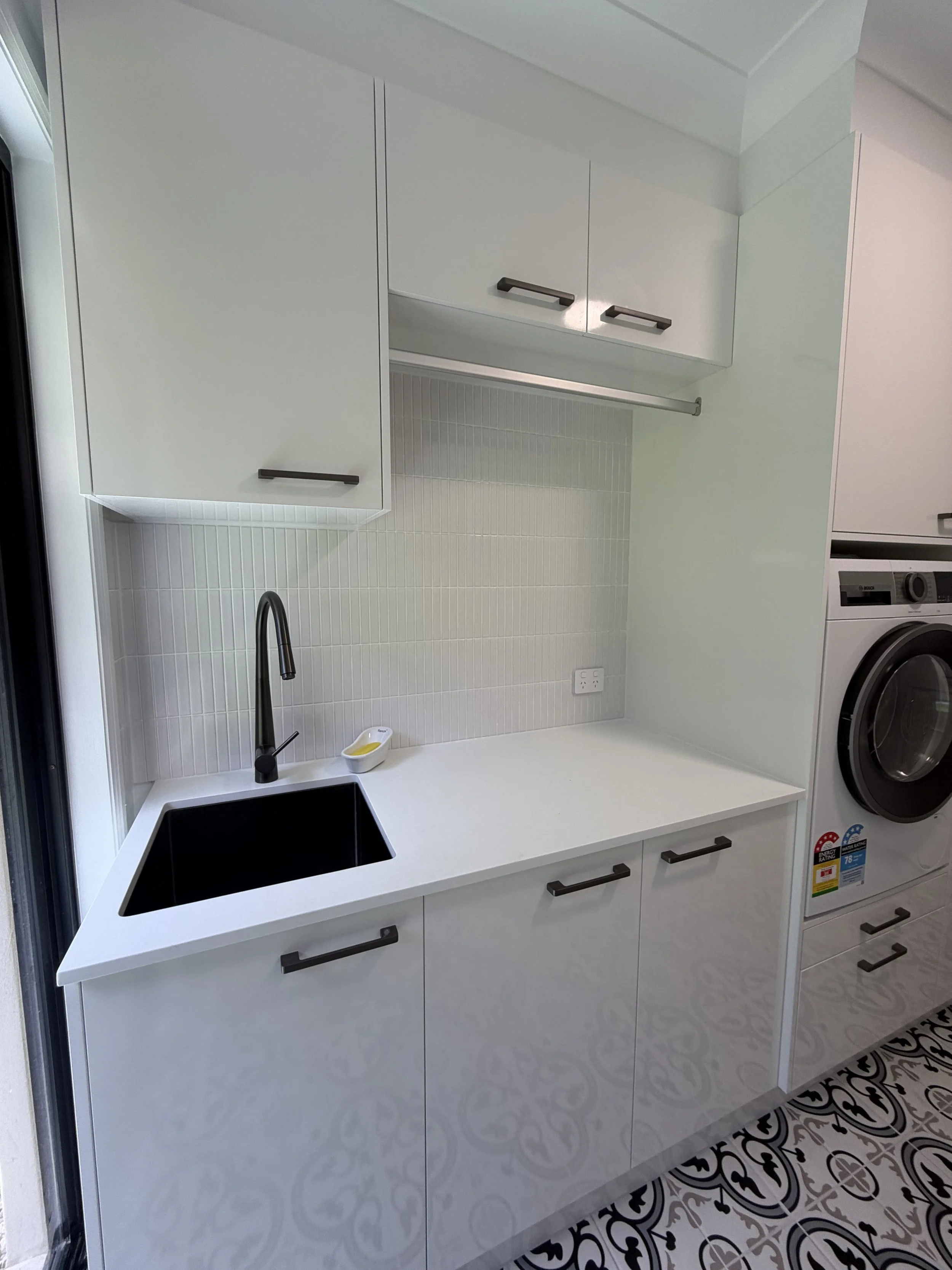 Modern laundry area with white cabinets, a black sink, a black faucet, a small soap dish, and stacked washer and dryer, with patterned tile flooring.