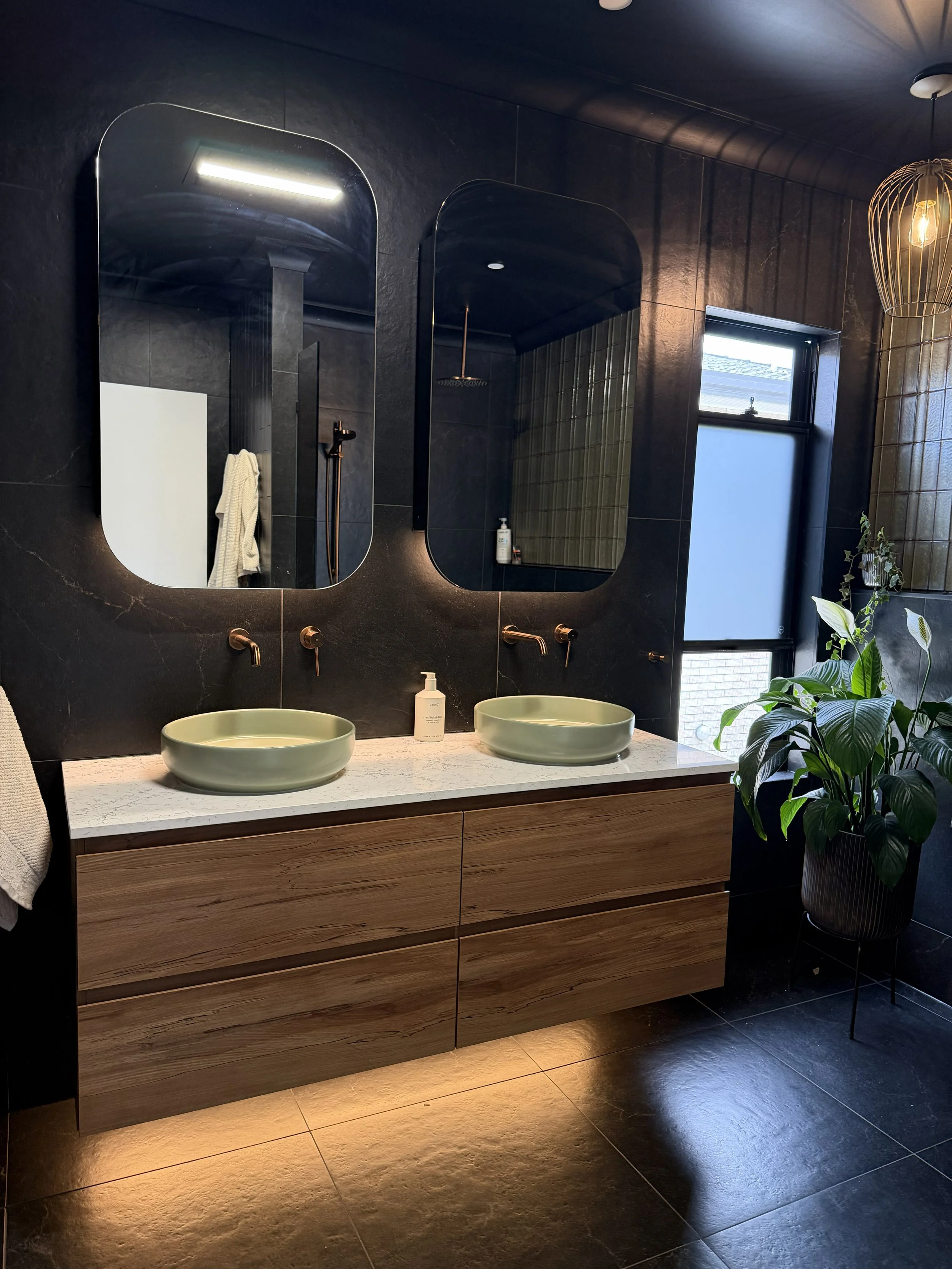 Modern bathroom with black tiles, a wooden vanity with a white marble countertop, two green vessel sinks, wall-mounted copper faucets, two oval mirrors, a large window, a potted plant, and a hanging light fixture.