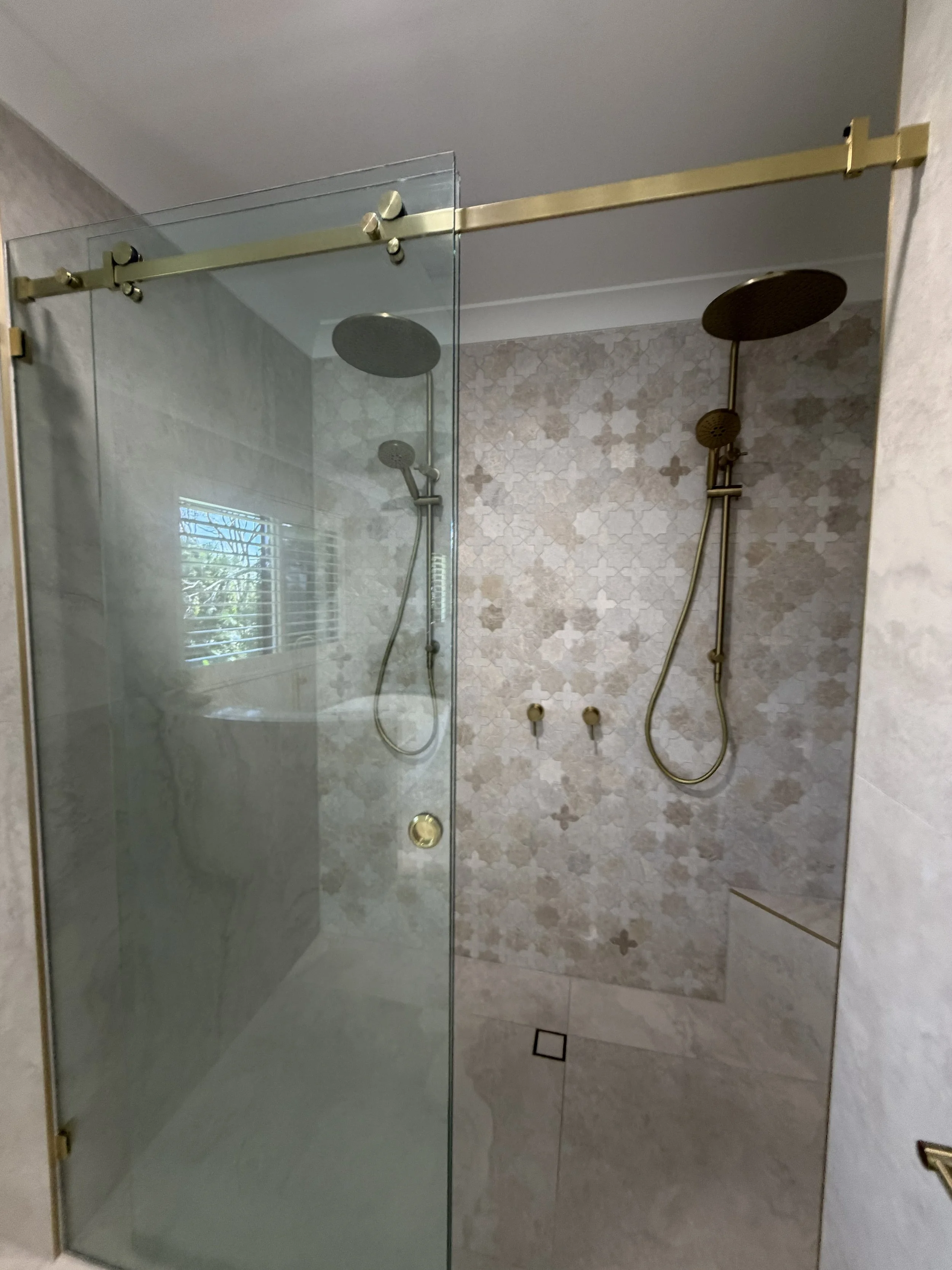 Modern shower with a glass door, featuring dual rainfall showerheads and handheld shower fixtures, set against a wall with geometric patterned tiles and neutral-colored marble tiles.