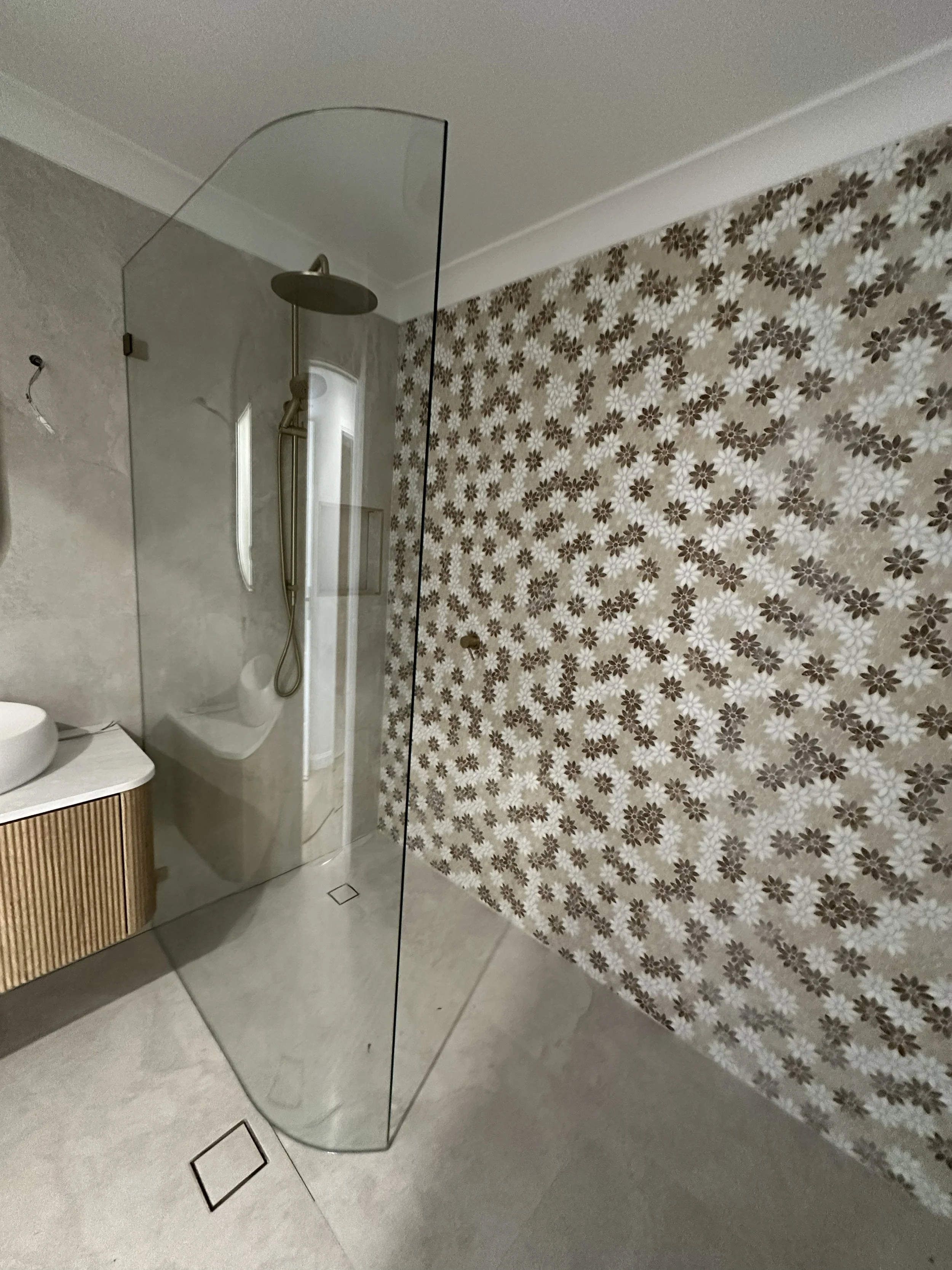 Bathroom with a walk-in shower enclosed by glass, a patterned floral wallpapered wall, a wooden cabinet with a white vessel sink, and a showerhead with a handheld attachment.