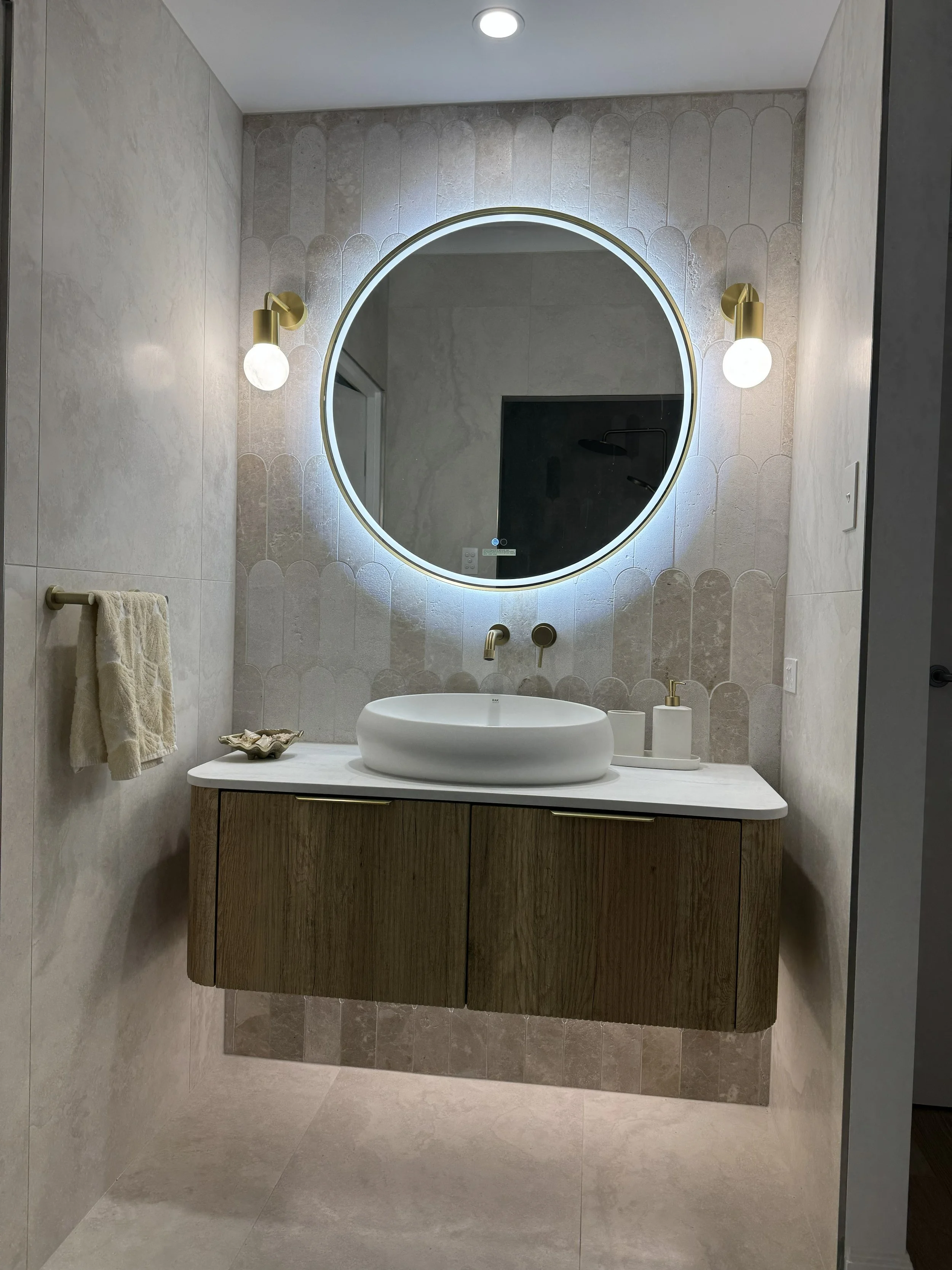 Modern bathroom with a round backlit mirror, dual brass wall sconces, a vessel sink on a wooden vanity, and minimal decor.