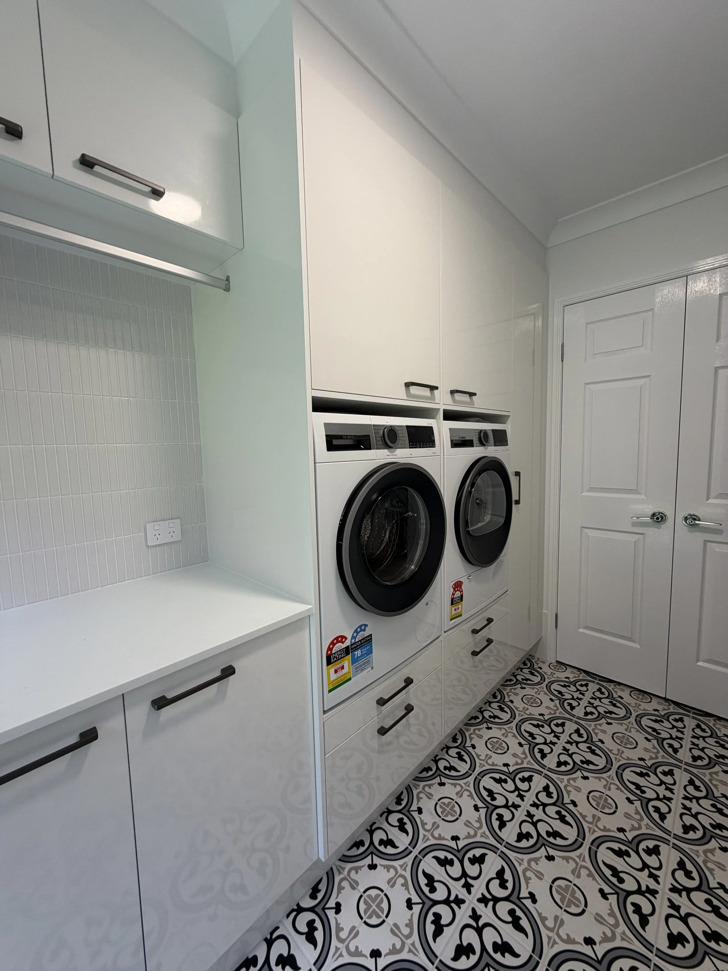 Modern laundry room with white cabinets, a front-loading washer and dryer, black handles, decorative floor tiles, and a door.