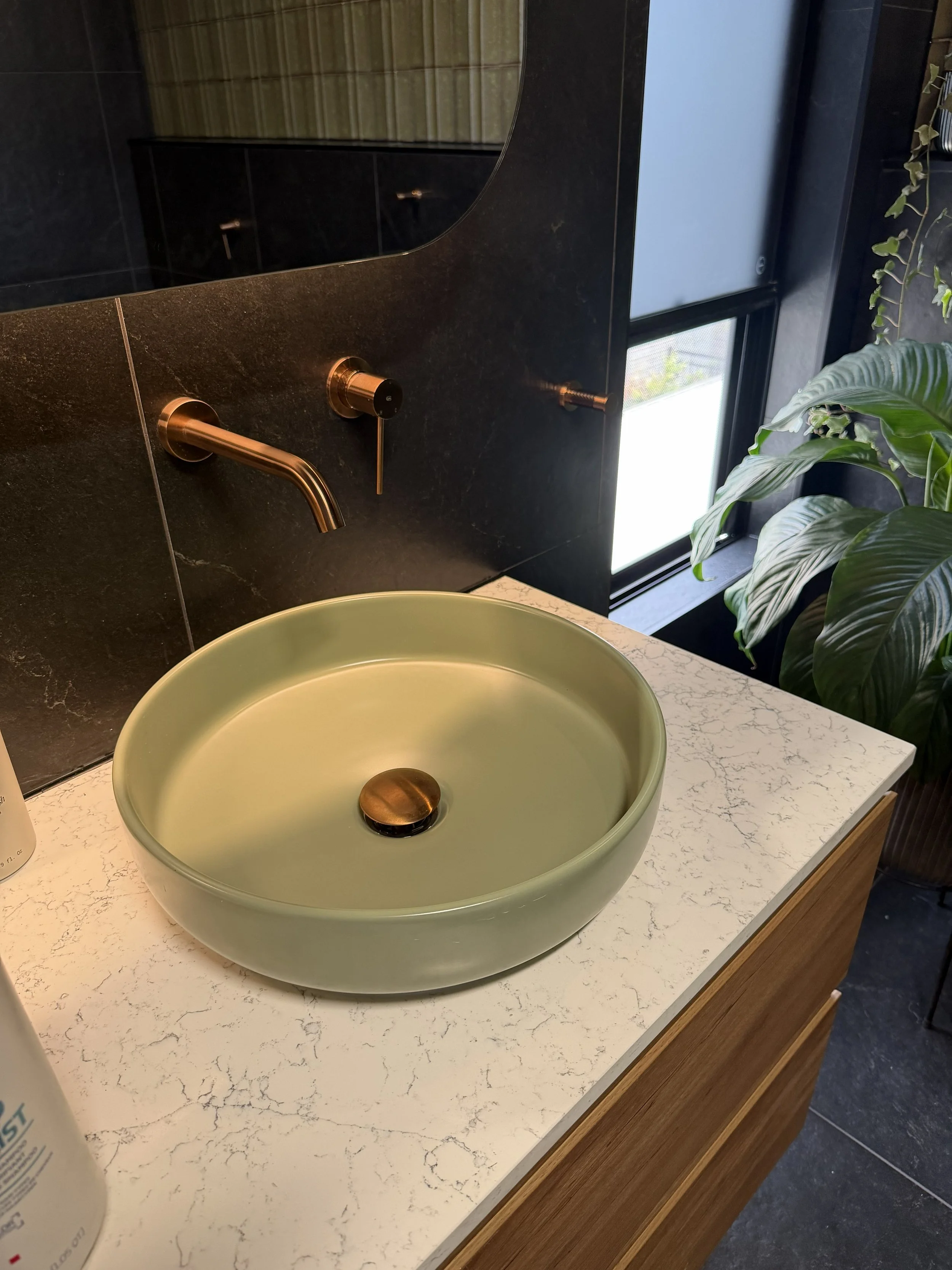 A modern bathroom sink area with a round, light green vessel sink, a wall-mounted faucet, and a large mirror. There is a window and some greenery nearby.
