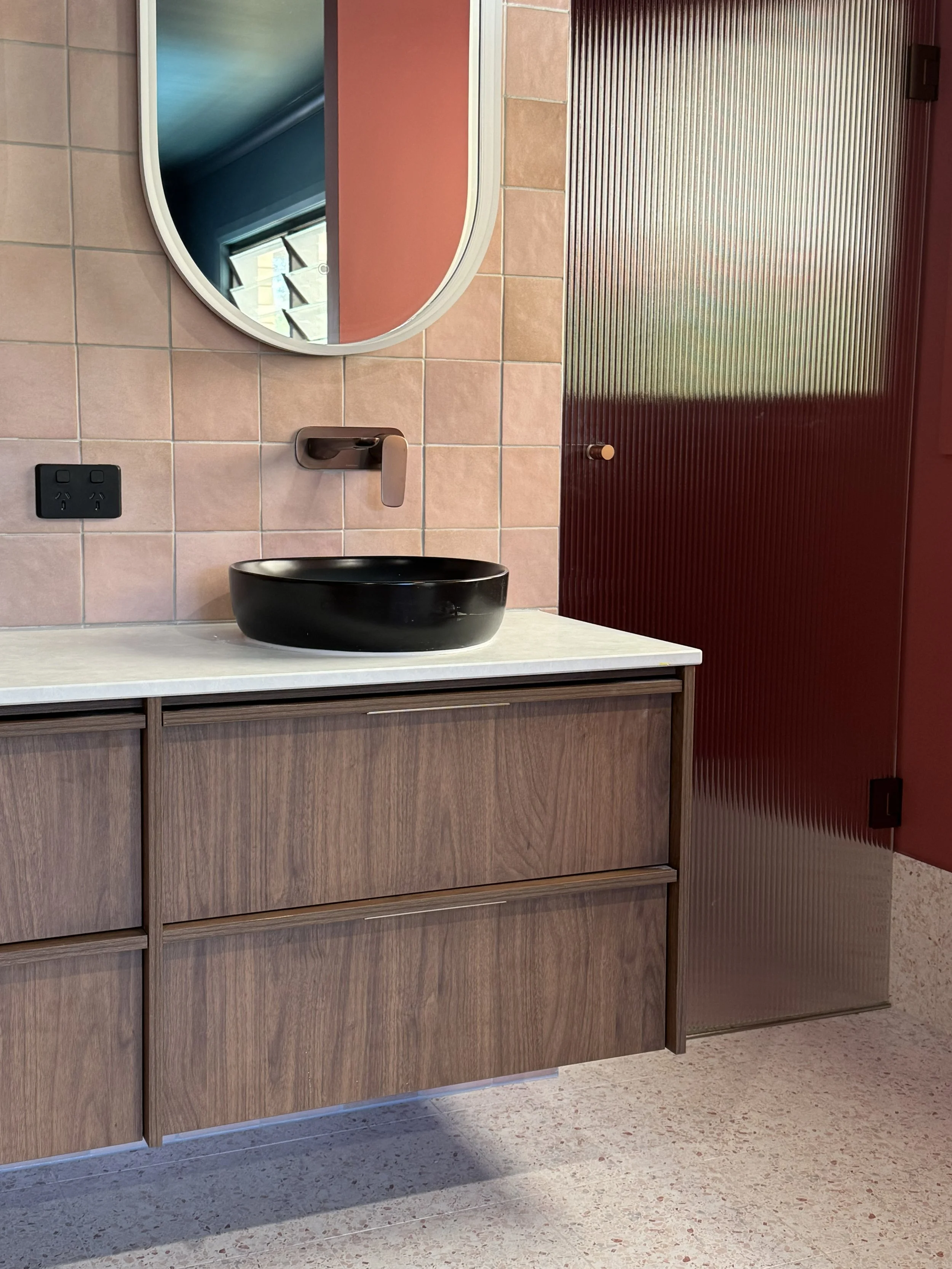 Modern bathroom vanity with a black vessel sink, a wall-mounted faucet, a round mirror, pink tiled wall, a red textured door, and a power outlet.