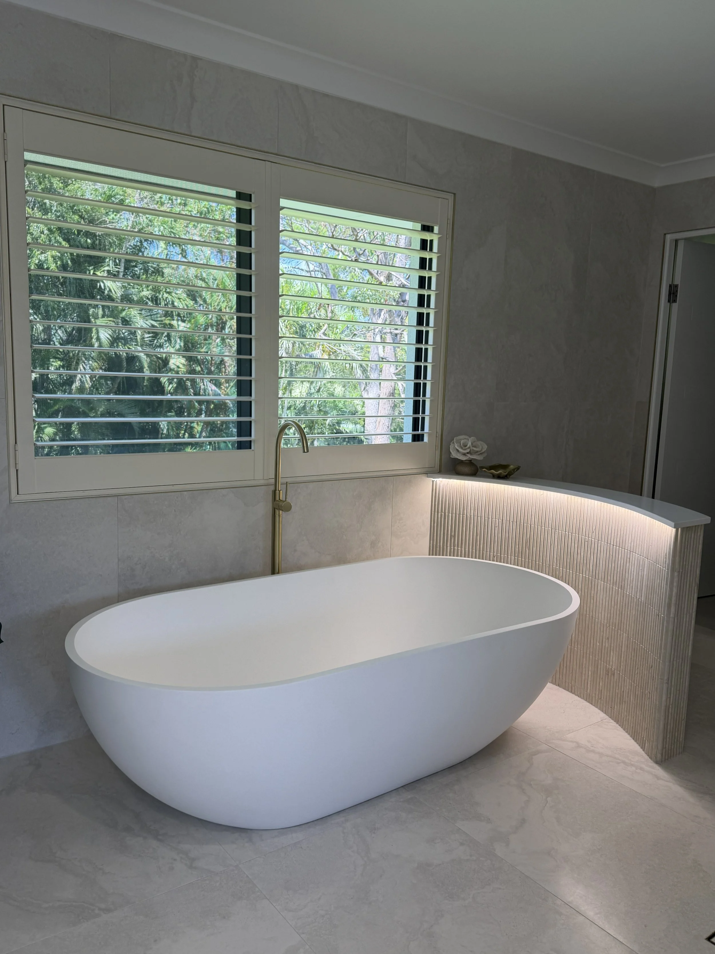 Modern bathroom with a freestanding white bathtub, a large window with plantation shutters, and a curved vanity with lighting, decorated with a small flower arrangement and a bowl.