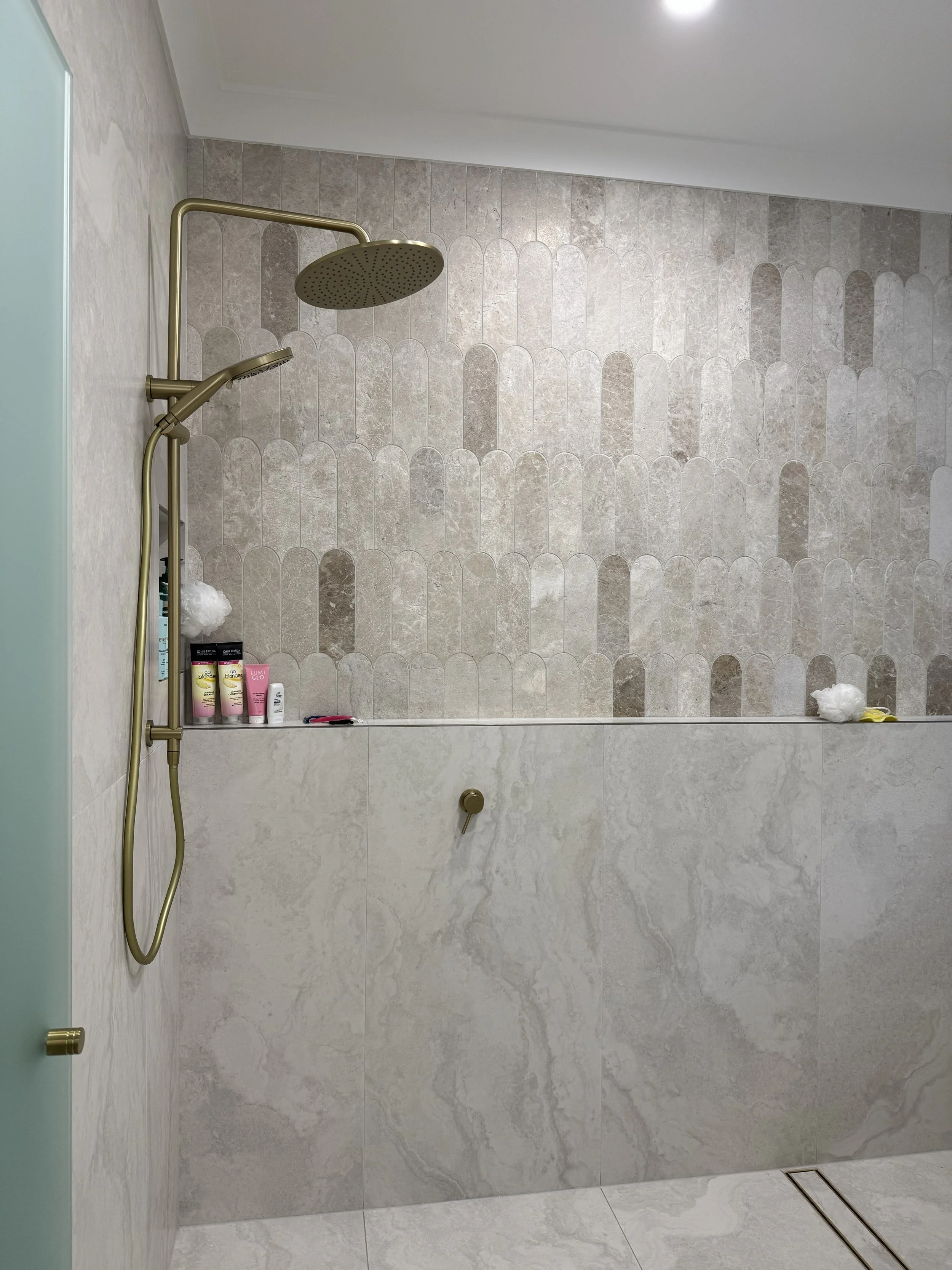 An empty walk-in shower with beige and gray tiled walls, including a large overhead rainfall showerhead and a handheld showerhead, with bathroom products on a small shelf.